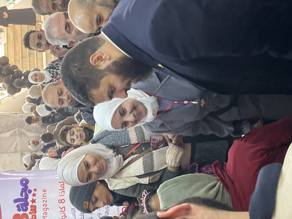 An indoor scene in Kafr Batna, Syria, depicts a crowded event or gathering. In the immediate foreground, a man with a dark beard, wearing a dark blue suit jacket over a light collared shirt, is seen in profile on the right, looking down and to his left with a smile. Adjacent to him, a woman wearing a white hijab and a grey coat with a red lanyard and a dark identification badge around her neck is smiling and looking towards the bearded man. To her left, another woman with a beige hijab, a grey vest, a beige and striped scarf, and light-colored fingerless gloves is smiling and looking downwards. A small brown cross-body bag is visible across her chest. A young boy in a dark beanie is positioned to her left, looking down. In the mid-ground, a diverse crowd of individuals, including several men and women in hijabs, is present. A young boy with dark markings on his cheek, wearing a yellow jacket, is visible. Another woman in a white hijab and a black and white plaid shirt stands further back. In the background, a large white banner with red Arabic text, including the word "Magazine" and "8 كانون" (8 January), is visible on the left. To the right, a set of light-colored stairs adorned with black balloons ascends. The overall scene suggests a public event with positive interactions among attendees.