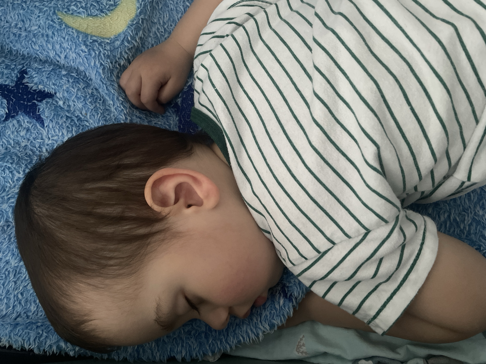 A toddler is sleeping on a blue blanket with a moon and star pattern. The child is wearing a white shirt with green stripes. The child has brown hair. No information is present to identify this picture’s location in Izhevsk, Russia.