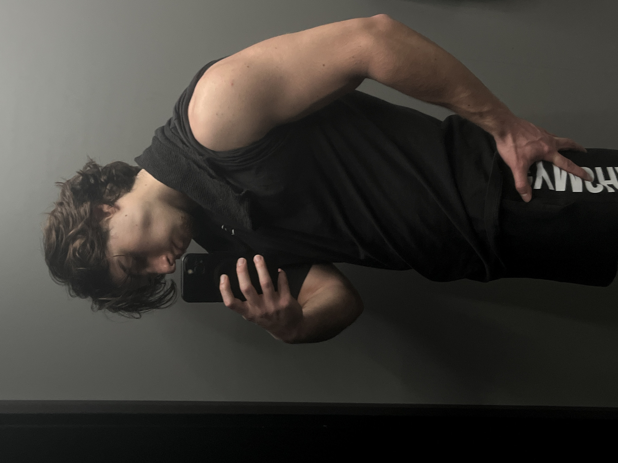 A person is taking a selfie in front of a mirror. The person has short, dark hair and is wearing a black tank top and black athletic pants. The person is holding a black cell phone in their right hand and looking at the phone's screen. The background is a gray wall and a black baseboard. The location is Unknown, Unknown.