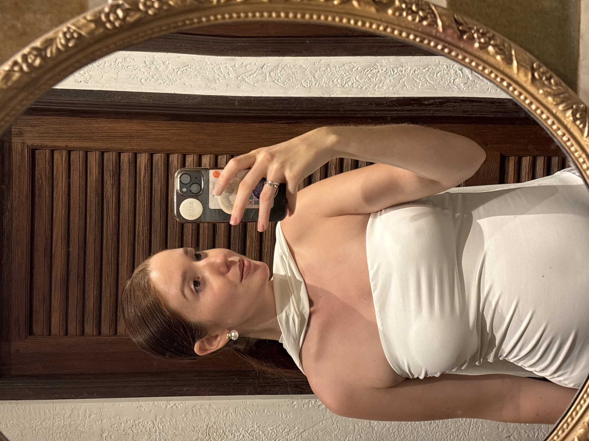 A woman is taking a selfie in a mirror. The mirror has a gold, ornate frame and reflects a wooden door with vertical slats and a white textured wall above it. The woman is wearing a white dress. She is holding her phone in her right hand, and the phone has stickers on the back. The earring she is wearing is a pearl. The location cannot be determined from the image.
