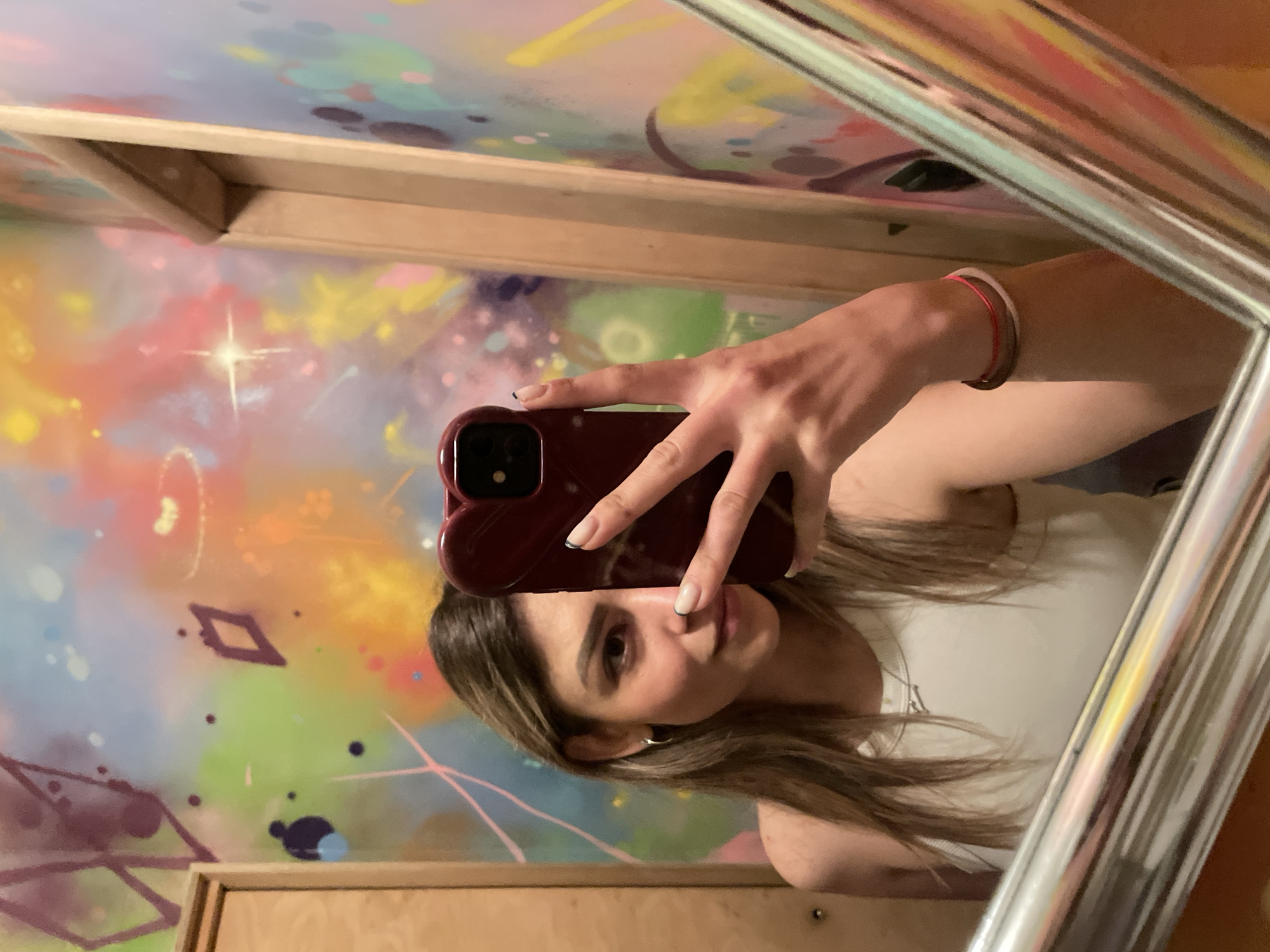 An individual is captured in a mirror selfie within an indoor setting in the city of Unknown, Unknown. The subject is a female with long, light brown hair and subtle makeup, wearing a white ribbed tank top. She has a silver hoop earring in her visible left ear and a thin silver necklace. On her right wrist, two thin bracelets are present: one dark and one pink. Her fingernails exhibit a dark French tip design. She holds a smartphone with a dark red, heart-shaped case, covering the right side of her face. The phone's camera lenses are visible on the case. The background wall is vibrantly painted with abstract, graffiti-style artwork featuring splatters, lines, and shapes in various colors including pink, yellow, orange, green, blue, and purple. Distinct patterns on the wall include diamond shapes, a prominent starburst effect, and a faint smiley-face-like figure. Wooden door frames are visible on the left and right sides of the reflected image, flanking the mirror. A reflective, metallic bar or frame runs along the bottom edge of the mirror.
