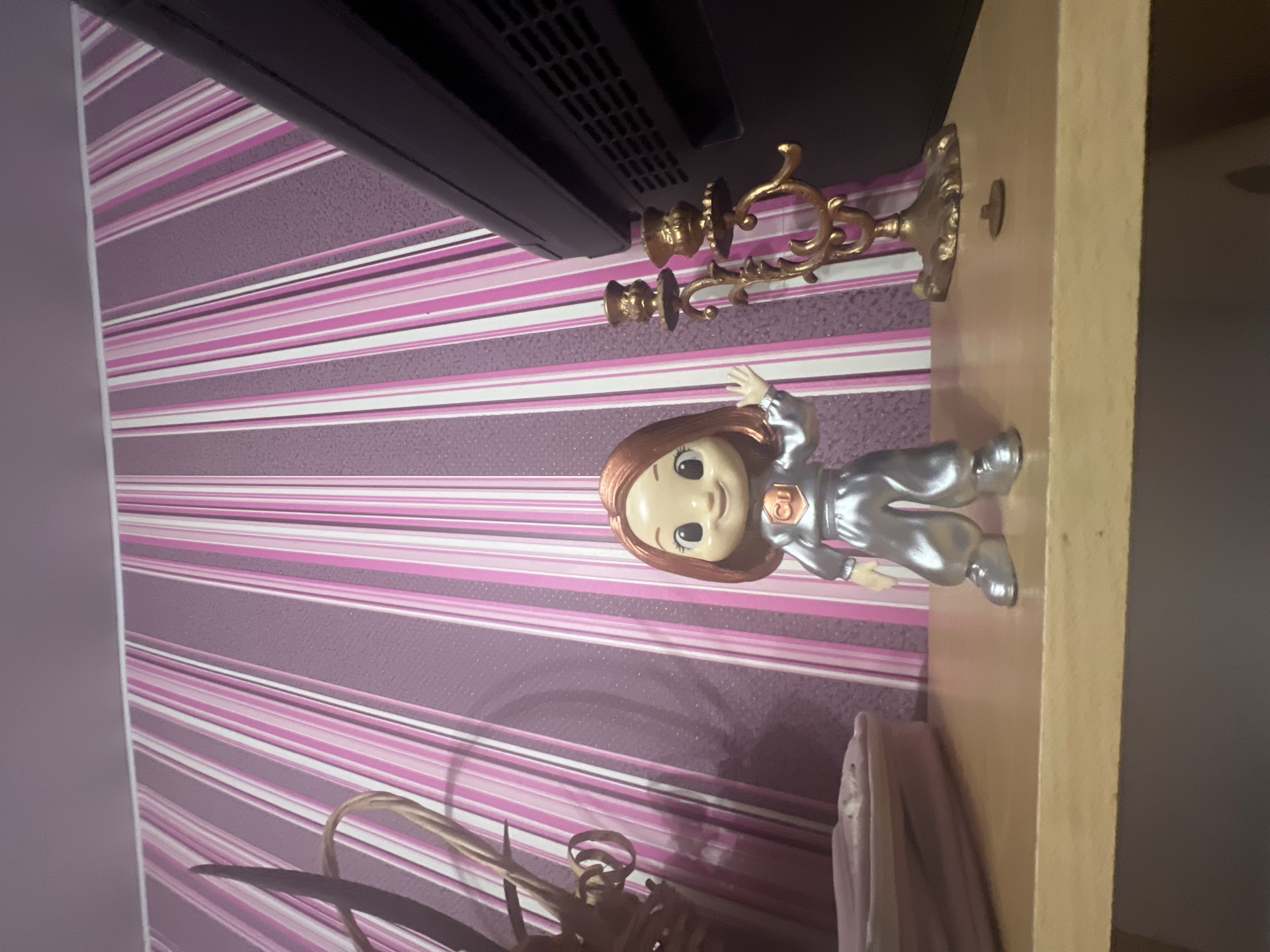 An action figure of a young girl with red hair, wearing a silver jumpsuit with a copper symbol on the chest, stands on a light-colored wooden shelf. The figure has its left hand raised in a wave. To the right of the figure, a golden candelabra with two candle holders stands on the shelf. In the background, a wall is covered with vertical stripes of pink and purple. To the left of the figure, a woven basket with decorative elements is visible, and further to the left, a pink rectangular object, possibly a case or bag, rests on the shelf. A dark object, likely a television or monitor, is positioned on the right side of the shelf, partially obscuring the background. A small, dark, circular object is on the shelf in front of the candelabra. The image appears to be taken indoors, on a shelf that is part of a room's decor. The location is not specified within the image, and therefore cannot be determined to be Saratov, Russia.