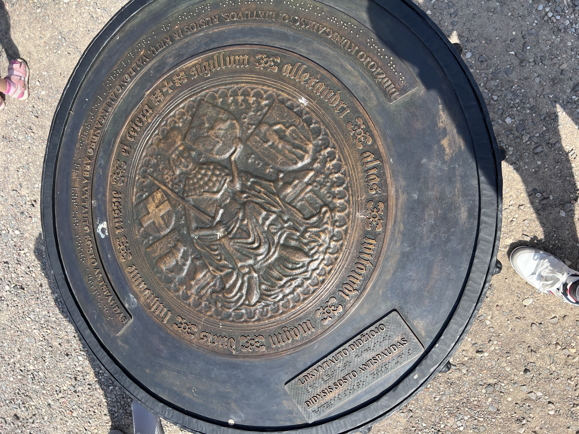 This is a medium, high-angle, outdoor, close-up, full shot of a large, circular, bronze plaque embedded in the ground, likely made of gravel. The plaque features an embossed central design of a knight on horseback, possibly Saint George slaying a dragon, surrounded by a decorative border with what appears to be Lithuanian text and Braille. Some of the text is partially obscured by shadow. On the left side of the plaque, a separate rectangular plaque with more text and Braille is affixed. The surrounding ground is covered in small, light-colored gravel and pebbles. In the upper right corner, the feet of a child wearing pink sandals are visible. In the lower portion of the image, a white sneaker with red and blue accents is positioned near the plaque. The overall lighting suggests a sunny day, with distinct shadows cast by the plaque and the surrounding objects. This plaque is located at the site of the Trakai Castle in Trakai, Lithuania, marking the "Footprint of Vytautas the Great."
