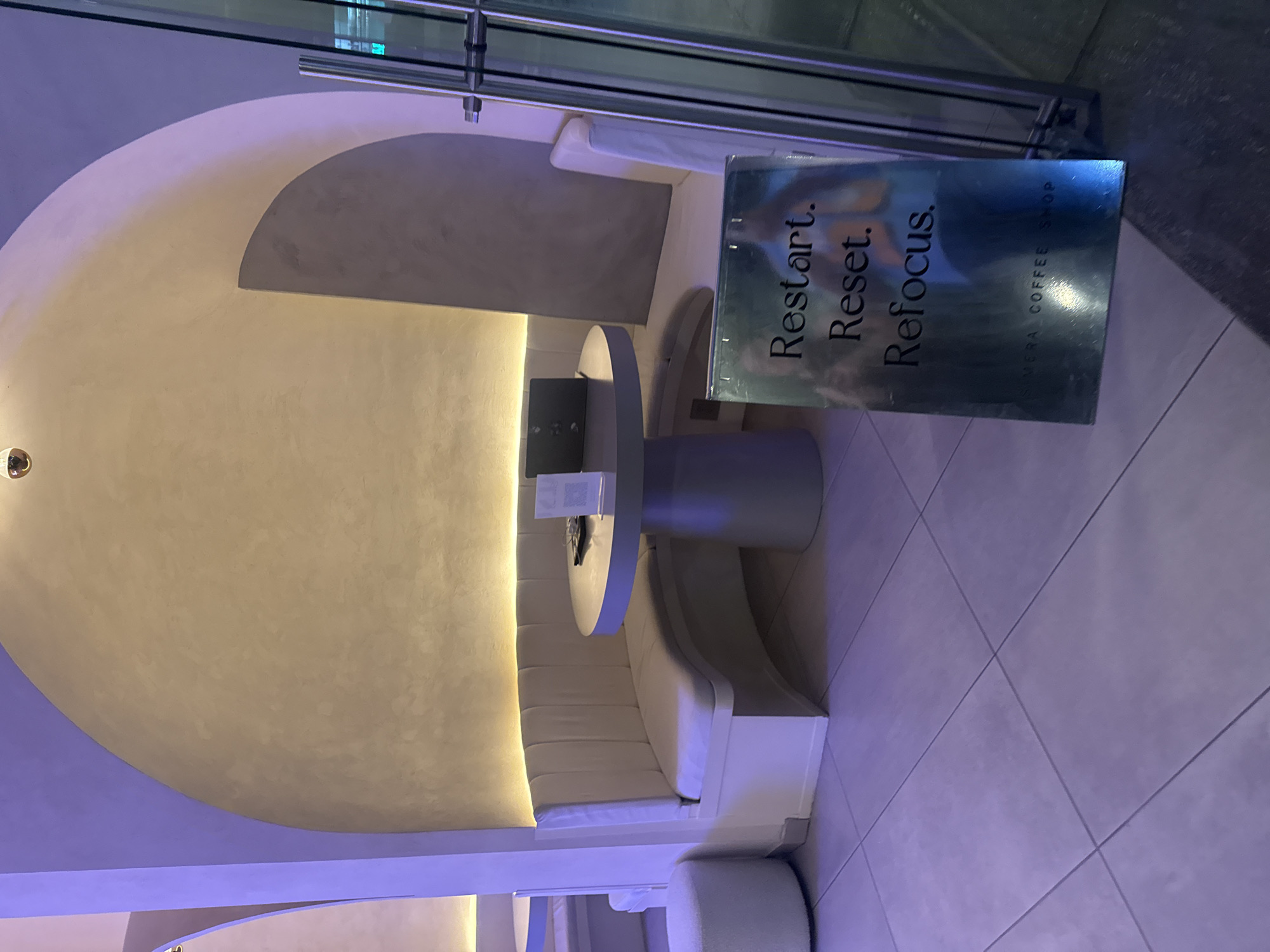 A booth with curved banquette seating surrounds a cylindrical table in a modern interior space, likely a coffee shop based on a sign in the foreground that reads "Restart. Reset. Refocus. SIMORA COFFEE SHOP." The space features recessed lighting along the wall and a unique architectural design with a partially enclosed dome-like ceiling. The floor appears to be tiled. The precise location is the city of San Bernardino Tlaxcalancingo, Mexico. No people are directly visible in the image, but reflections on the sign suggest their presence.