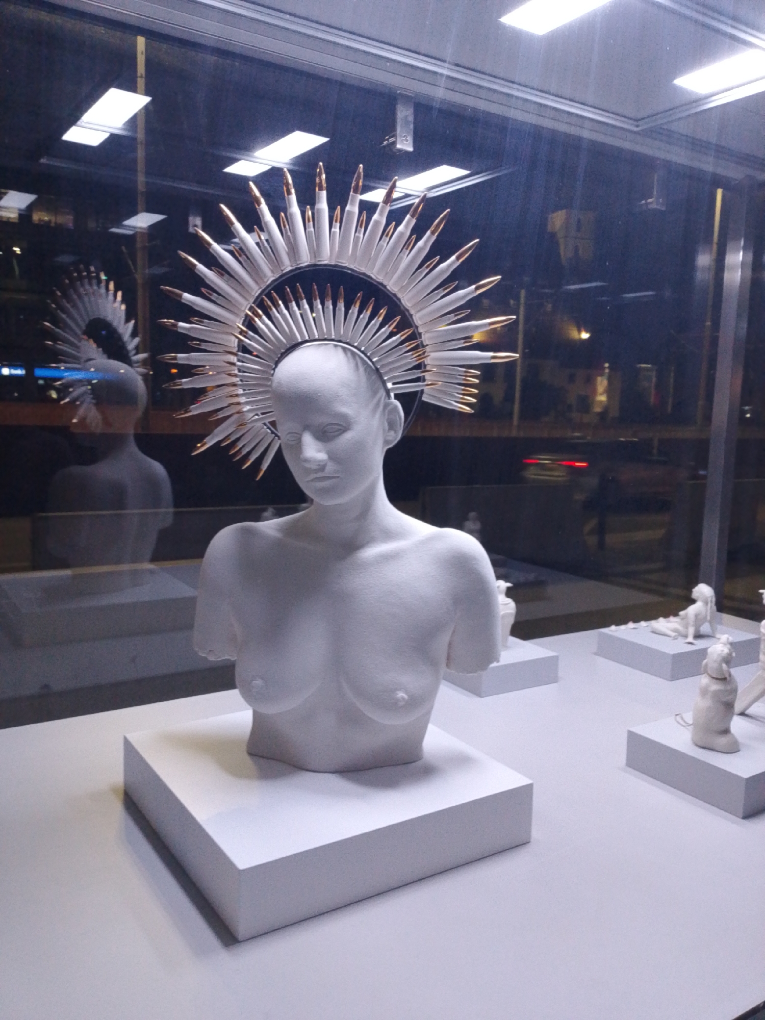 The image shows a white, headless bust of a person with breasts, wearing a halo made of what appear to be bullets. The bust is displayed in what looks like a museum or gallery window, with reflections of lights and city buildings visible in the glass. The background shows a street scene at night with some cars and buildings. The bust is placed on a white pedestal. The overall impression is one of a contemporary art installation. The location is the National Museum in Wrocław, Poland.