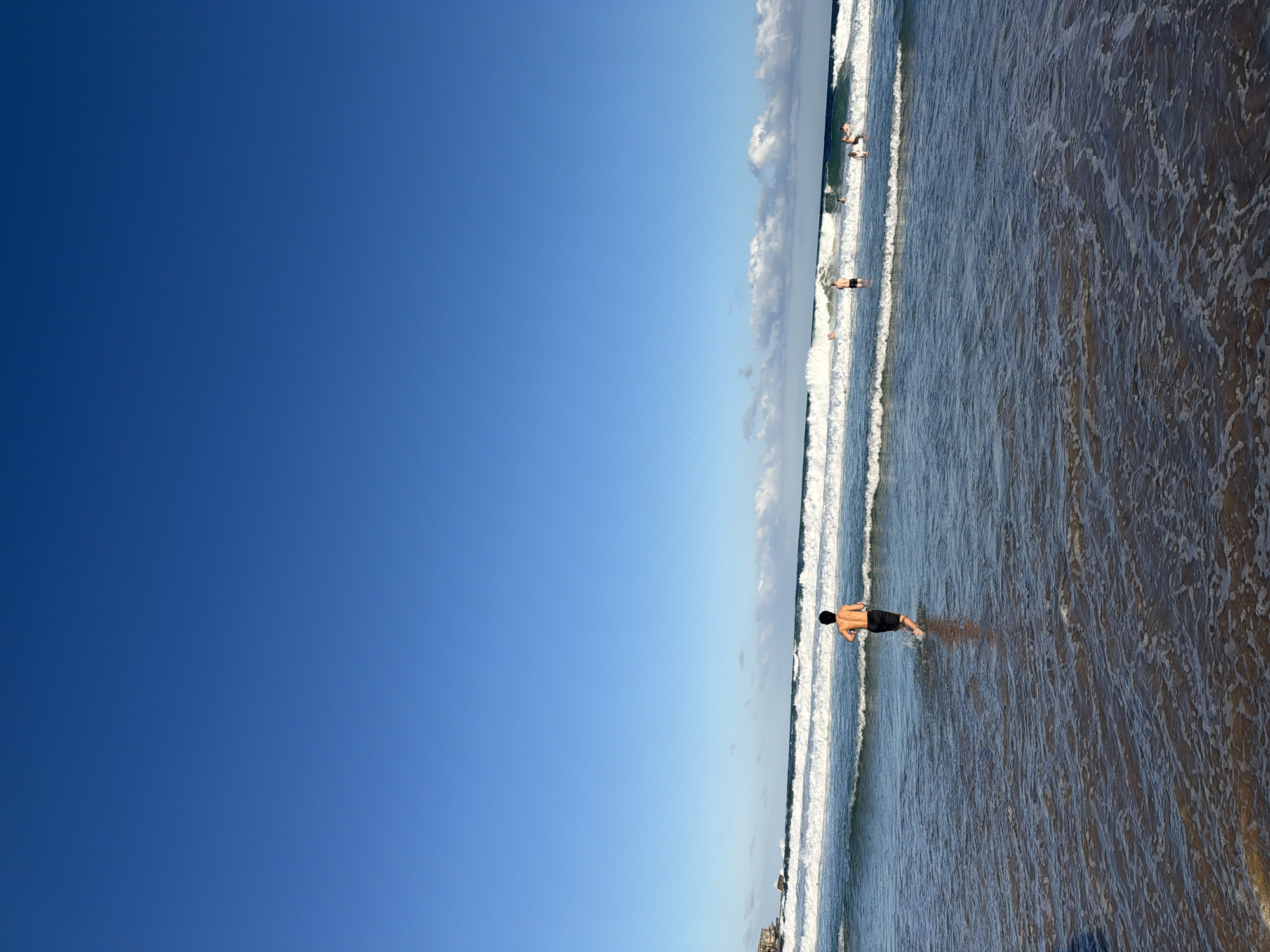 At Bondi Beach, Australia, under a clear blue sky, a young man runs into the ocean, his back to the camera. He wears black swim trunks. Multiple other people are visible in the water, some swimming, others standing, further out in the ocean. The waves are breaking and rolling towards the shore, creating a dynamic scene. The sand is wet and shallow water covers the foreground. In the distance, cliffs can be seen on the left side of the frame.