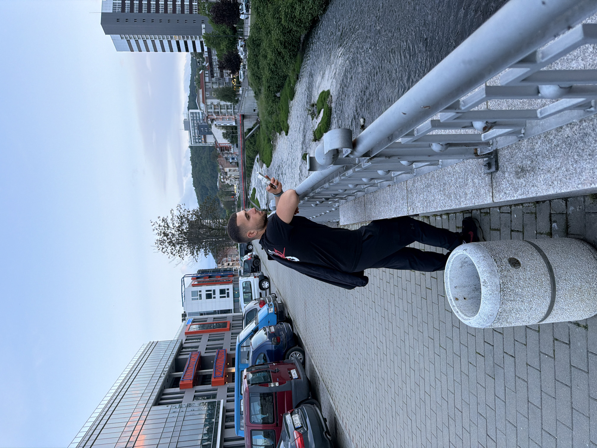 A man is photographed in the city of Gabrovo, Bulgaria. He is standing on a sidewalk near a bridge, leaning against the railing, and holding a smartphone. The bridge overlooks a river. The man is wearing a black shirt and pants, with a black jacket draped over his shoulder. Cars are parked on the other side of the sidewalk near buildings. There is a tall building and green trees visible in the background, along with other buildings and a sky with light clouds. The sidewalk is made of grey bricks, and a cylindrical concrete waste bin is nearby.