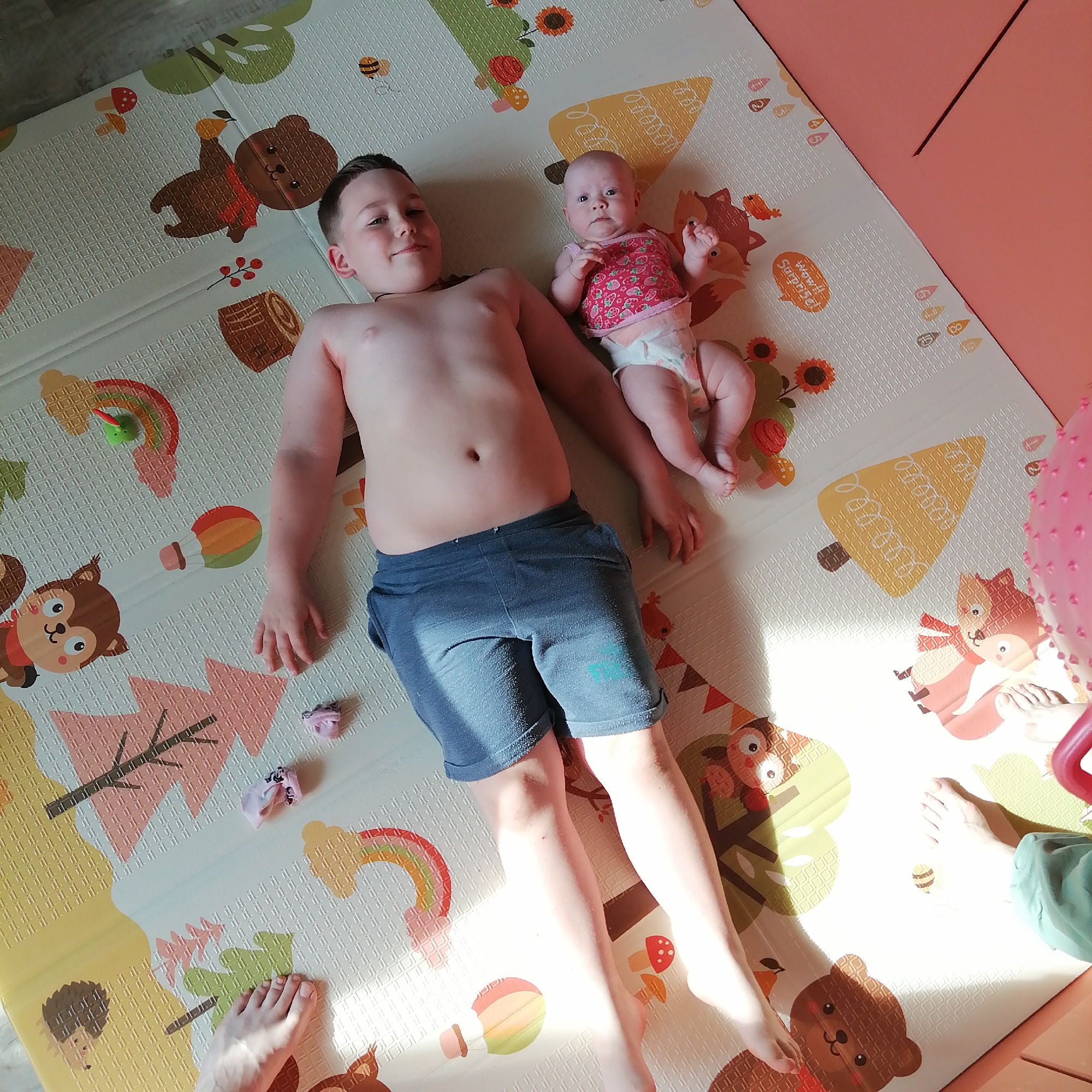 In this overhead shot, a young boy and a baby are lying on a colorful play mat patterned with cartoon forest animals and autumn leaves. The boy, who appears to be around 8-10 years old, is shirtless and wearing blue denim shorts. He is lying on his back, with his head turned to the side, looking towards the camera with a slight smile. Next to him, the baby is also lying on her back, dressed in a pink and white strawberry-patterned top and a diaper. The baby's gaze is directed upwards. The play mat is placed on a floor that appears to be wood or laminate. Sunlight streams in from the right, casting distinct shadows across the mat and highlighting the subjects. A pink, textured object, possibly a toy, is visible on the right edge of the frame. Scattered on the mat are a few small items, including tiny pink socks and what looks like a small toy rainbow with a green creature on it. The overall scene suggests a casual, indoor play session on a bright day. The setting is likely a child's room or a living area within a home. No visible text is present in the image.