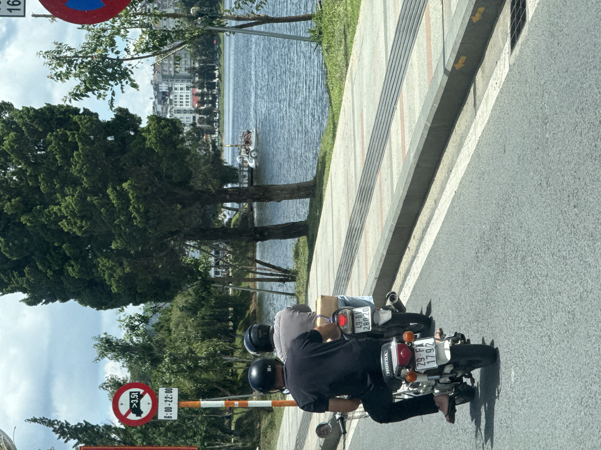 In Da Lat, Vietnam, two individuals ride on a single motorcycle along a paved road. The rider in the front, wearing a black helmet and black shirt, is driving. Behind him, a passenger with a black helmet and a grey shirt sits holding a cardboard box. The motorcycle has a license plate with the characters "29-FA 1792". They are traveling on a road next to a sidewalk lined with trees. In the background, a lake is visible with a swan-shaped paddle boat on it, and beyond that, buildings form the city skyline. A traffic sign indicating restrictions for vehicles over 3.5 tons from 6:00 to 22:00 is also present. The sky is partly cloudy.