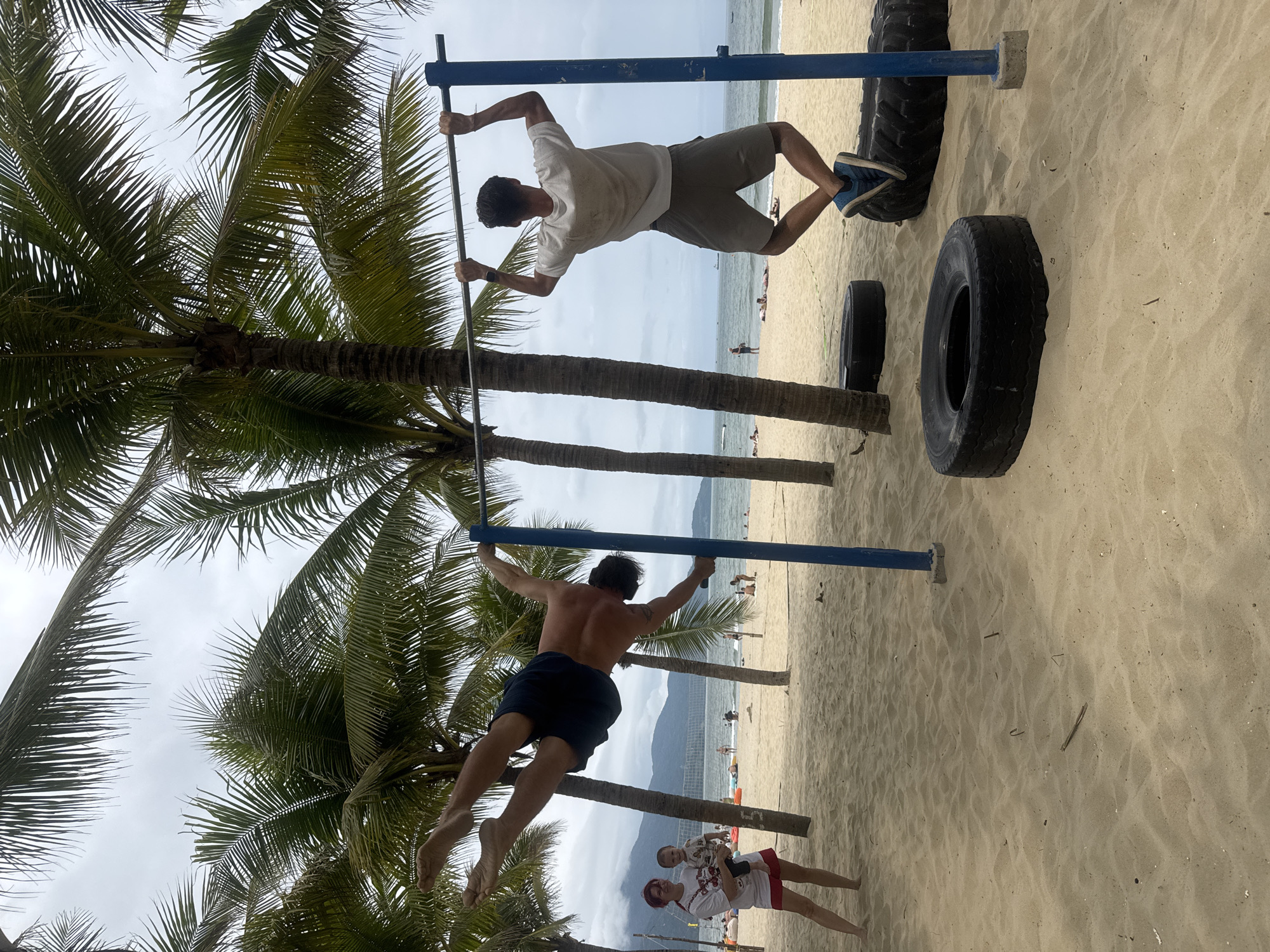Two men are hanging from parallel horizontal bars in an outdoor fitness area on a beach. The upper man wears a white T-shirt, gray shorts, and blue athletic shoes. The lower man is shirtless and wears dark blue shorts. Several large black tires are on the sandy ground nearby. Palm trees are around the workout area. Further down the beach, there are several people and the sea. Mountains are visible in the distance. The sky is overcast. In the lower right corner, two women and a baby are standing on the beach. The location is likely along My Khe Beach in Đà Nẵng, Vietnam, given the landscape and beach features.