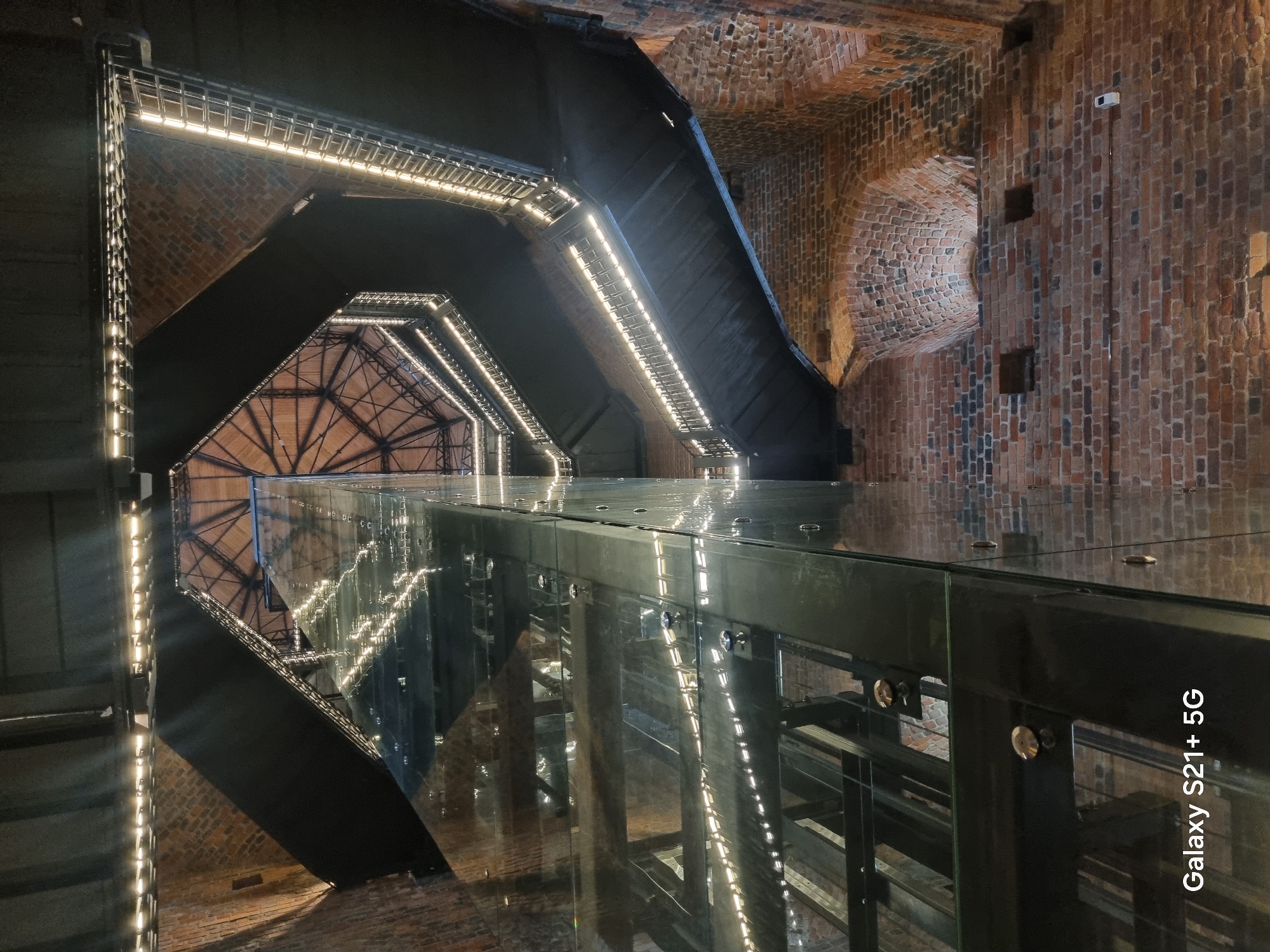 The image shows an interior view looking upwards inside a cylindrical brick tower. A modern glass elevator shaft runs vertically up the center of the tower. Surrounding the elevator shaft are several levels of dark, metal walkways and staircases that follow the circular shape of the tower. These walkways are illuminated with strip lighting along their edges. The original brickwork of the tower is visible on the exterior walls, with some arched openings and recesses. The ceiling of the tower, visible at the top, is a wooden, vaulted structure with metal support beams. The overall impression is a juxtaposition of historical architecture with modern design elements. This scene is located within Vyborg Castle in Vyborg, Russia.