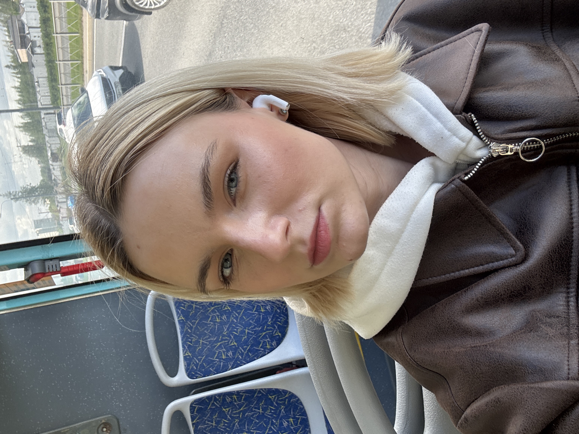 A fair-skinned person with blonde hair is seated on a bus. They are wearing a brown jacket over a white hooded garment and have a white wireless earbud in their left ear. The bus seats have a blue and white patterned fabric. Outside the bus window, a city landscape is visible, including buildings and vehicles, possibly in Saint Petersburg, Russia.