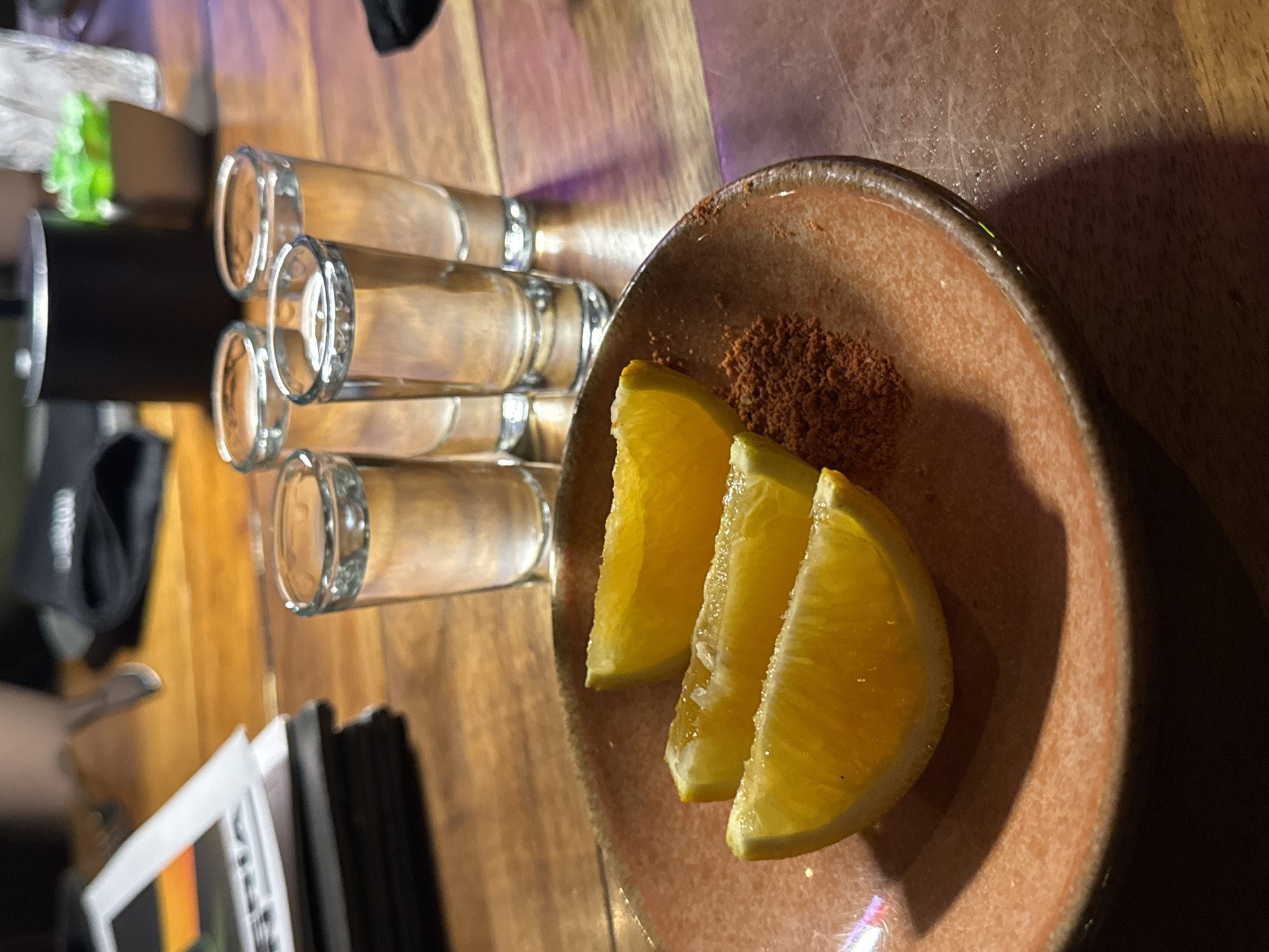 The image depicts a table in what appears to be a bar or restaurant in Puebla, Mexico, featuring a line of four shot glasses partially filled with a clear liquid, likely tequila or mezcal. Adjacent to the glasses is a small, circular, earthenware plate containing three orange slices and a mound of reddish-brown powder, which is likely ground worm salt or chili. The table itself is made of wood, with visible grain patterns and variations in color. Other items on the table include a black container with a green plant inside, a stack of printed materials, and a black cloth or napkin. The lighting is dim and creates shadows, suggesting an intimate or evening setting.