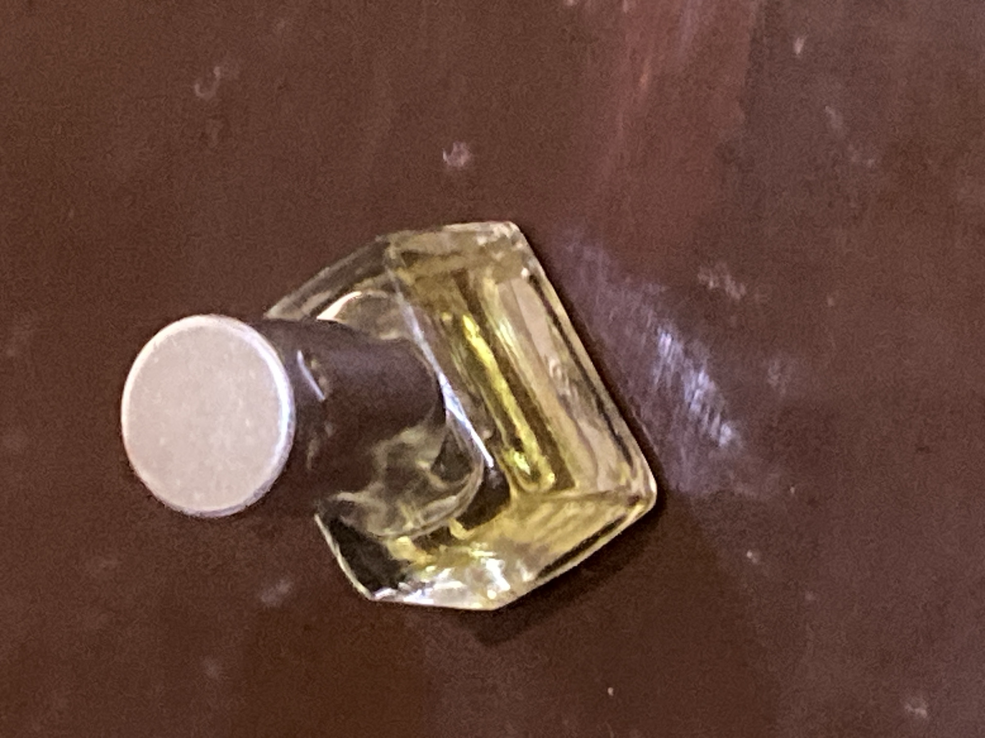 A close-up, high-angle shot shows a small, square, clear glass perfume bottle with a dark brown cap. The bottle is partially filled with a yellow liquid. The bottle rests on a dark brown, textured surface, possibly wood or laminate. The background is out of focus. The scene is located in Jauro Yinu, Nigeria.