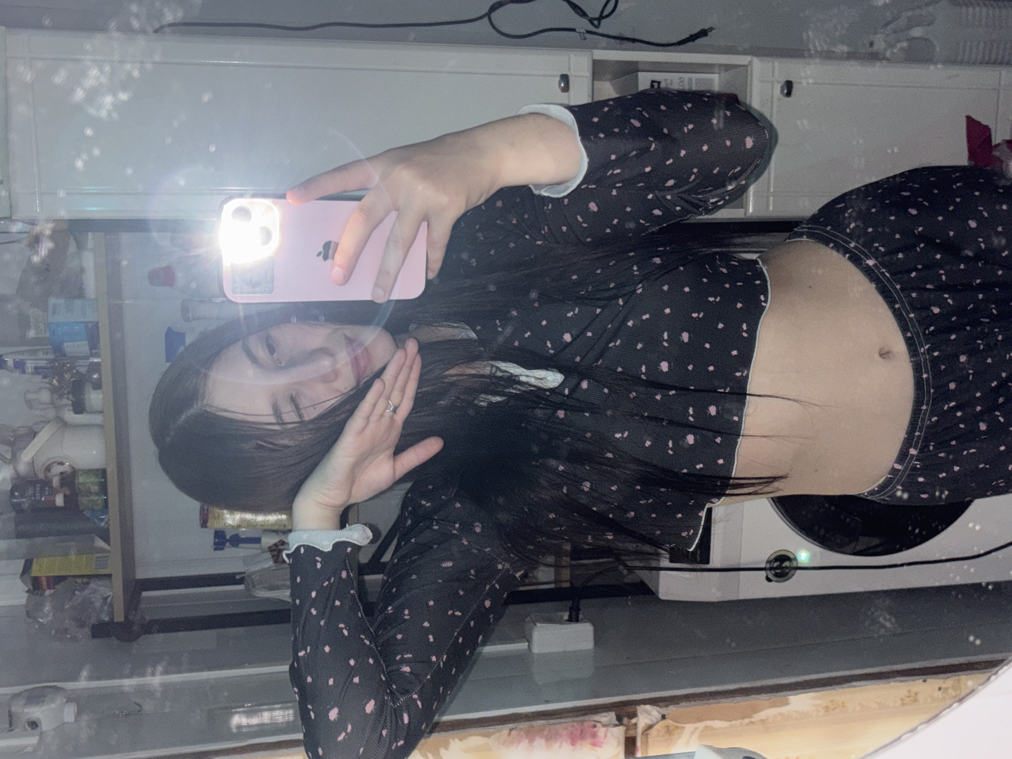 A young woman takes a mirror selfie indoors, likely in a kitchen or utility room in Фоки, Russia. She holds a pink smartphone with an active flash in her right hand, casting a bright light onto her face and the phone. Her face is partially visible, showing her left eye and mouth, framed by long, dark hair. She wears a dark, long-sleeved cropped top and matching pants, both adorned with small pink or light-colored polka dots. Her midriff and navel are exposed. A ring is visible on her left ring finger, and her left hand is raised near her face.
The background, reflected in the mirror, shows white cabinets or shelves above her, cluttered with various household items, bottles, and packages. Dark electrical wires are visible along the top edge of the reflection. To her right, a washing machine or similar appliance with a green indicator light is reflected. The ambient lighting appears dim, and numerous circular bokeh effects, possibly from reflections of light sources or dust on the mirror, are scattered across the image. No discernible text is clearly visible other than the iPhone logo on the phone case.
