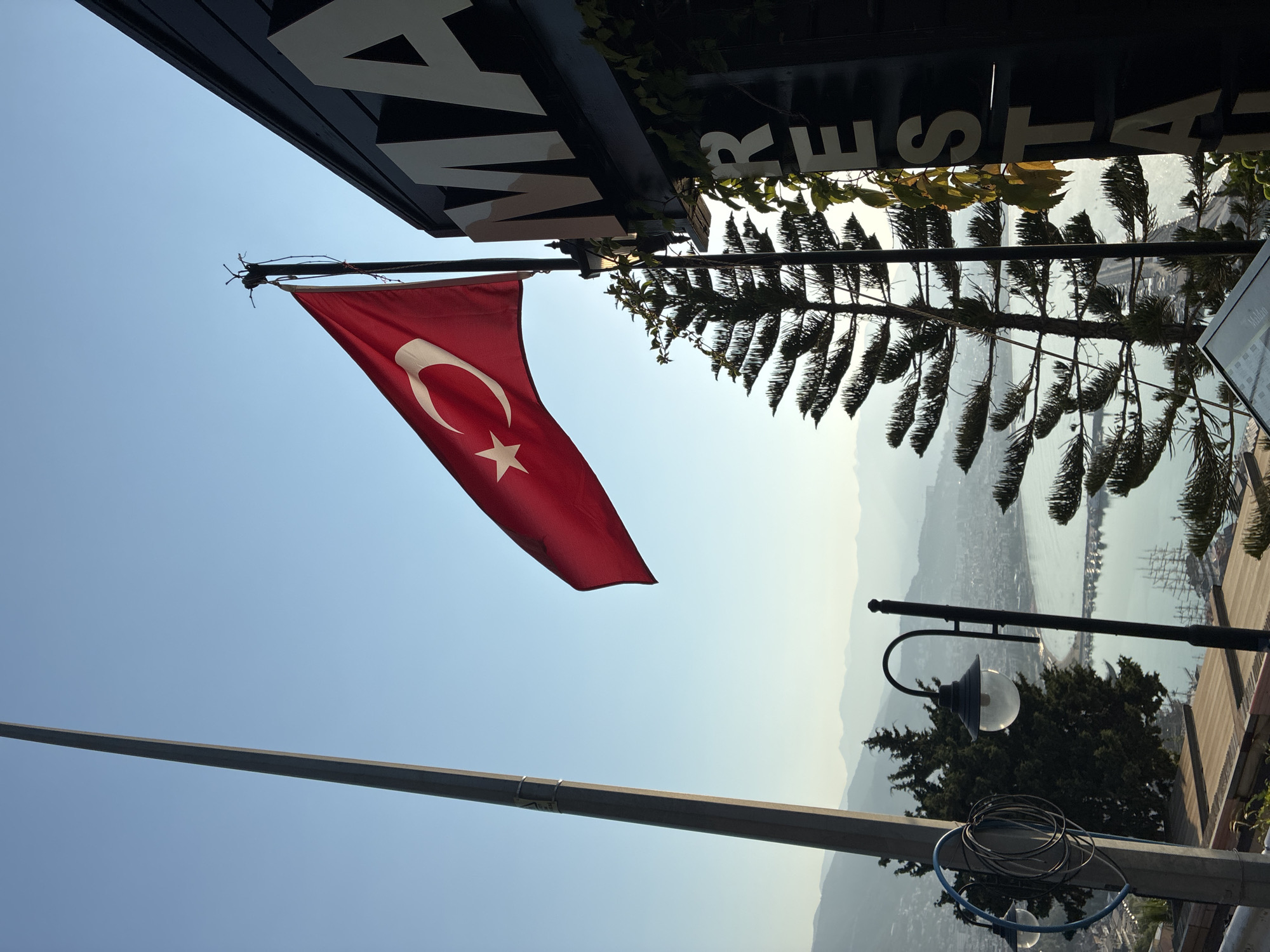 A low-angle, full shot captures a bright, clear blue sky above the city of Alanya, Turkey. In the foreground, a tall, thin metal pole stands on the left, a Turkish flag unfurls prominently from a flagpole on the right, its red fabric and white star and crescent silhouetted against the sky. To the right of the flag, a dark, rectangular building facade displays large, white, block letters that spell out "MA RESTAURANT". A tall, spiky evergreen tree grows in front of the building. The background reveals a hazy, sprawling cityscape with a body of water, possibly the Mediterranean Sea, dotted with sailboats, and a range of distant, mist-covered mountains. A streetlamp with a glass globe is visible in the middle ground.