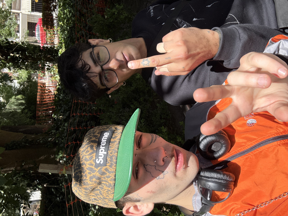 Two young men pose for a selfie outdoors. One wears a "Supreme" cap and an orange jacket with headphones around his neck; his face is tattooed. The other, wearing glasses and a black hoodie, raises his middle finger, displaying a hand tattoo. Both have piercings. Buildings and foliage form the blurred background. The scene is brightly lit, suggesting daytime.