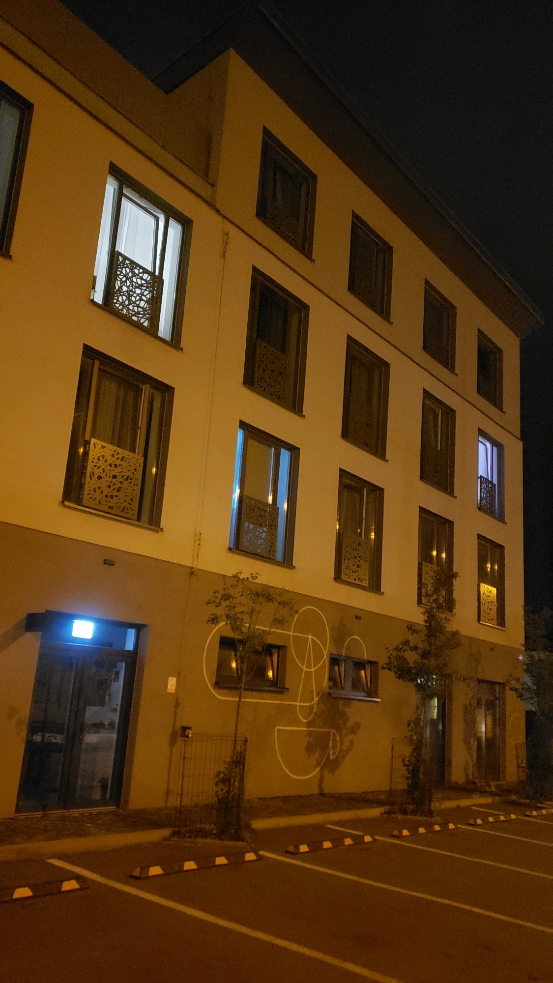 The image shows a multi-story building at night. The building is illuminated by warm yellow and some blue light emanating from the windows, some of which have decorative screens. The exterior walls are painted a light beige on the upper floors and a darker beige on the lower floor. On the lower floor, there is a glass-fronted entrance with a glowing blue sign above it. There is a stylized white graphic of a face and a cup on the wall of the lower floor. Two trees are planted in front of the building. The foreground shows a paved parking area with white parking lines and yellow and white concrete parking stops. The sky is dark. The building appears to be a hotel or guesthouse located in Prešov, Slovakia. There are no people visible in the image.