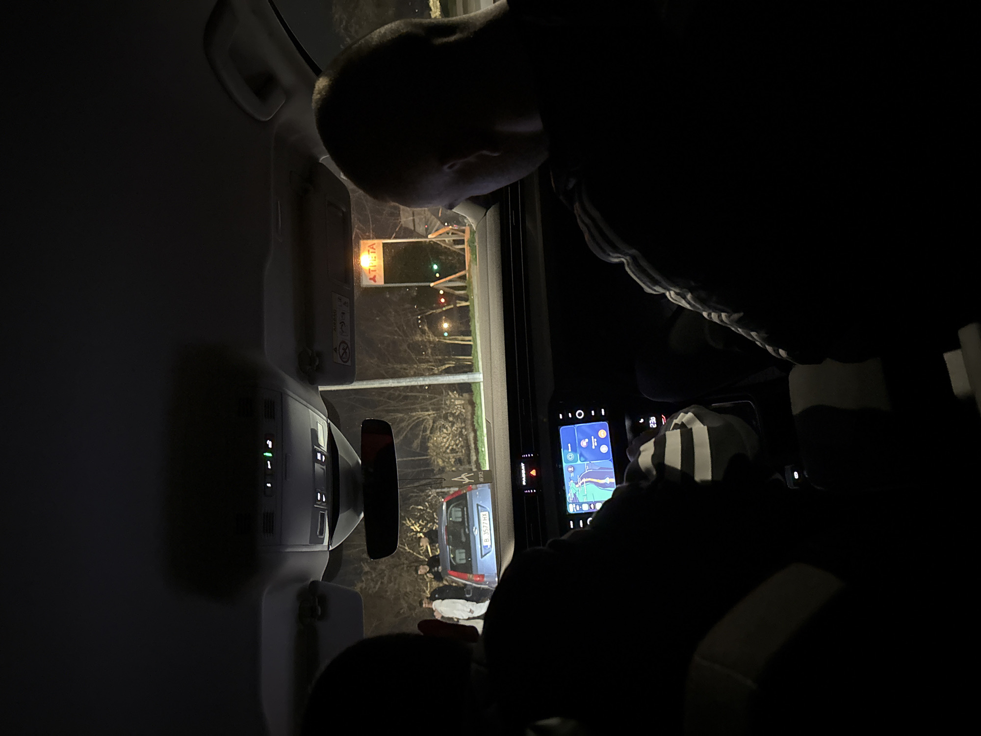 The image shows the interior of a car at night, with a person looking out the window. A navigation app is visible on the car's display. Outside, there are street lights and trees, as well as another vehicle with license plate "B 357 HX". The sign “ATLETA” is visible in the background. The location is Varna, Bulgaria.
