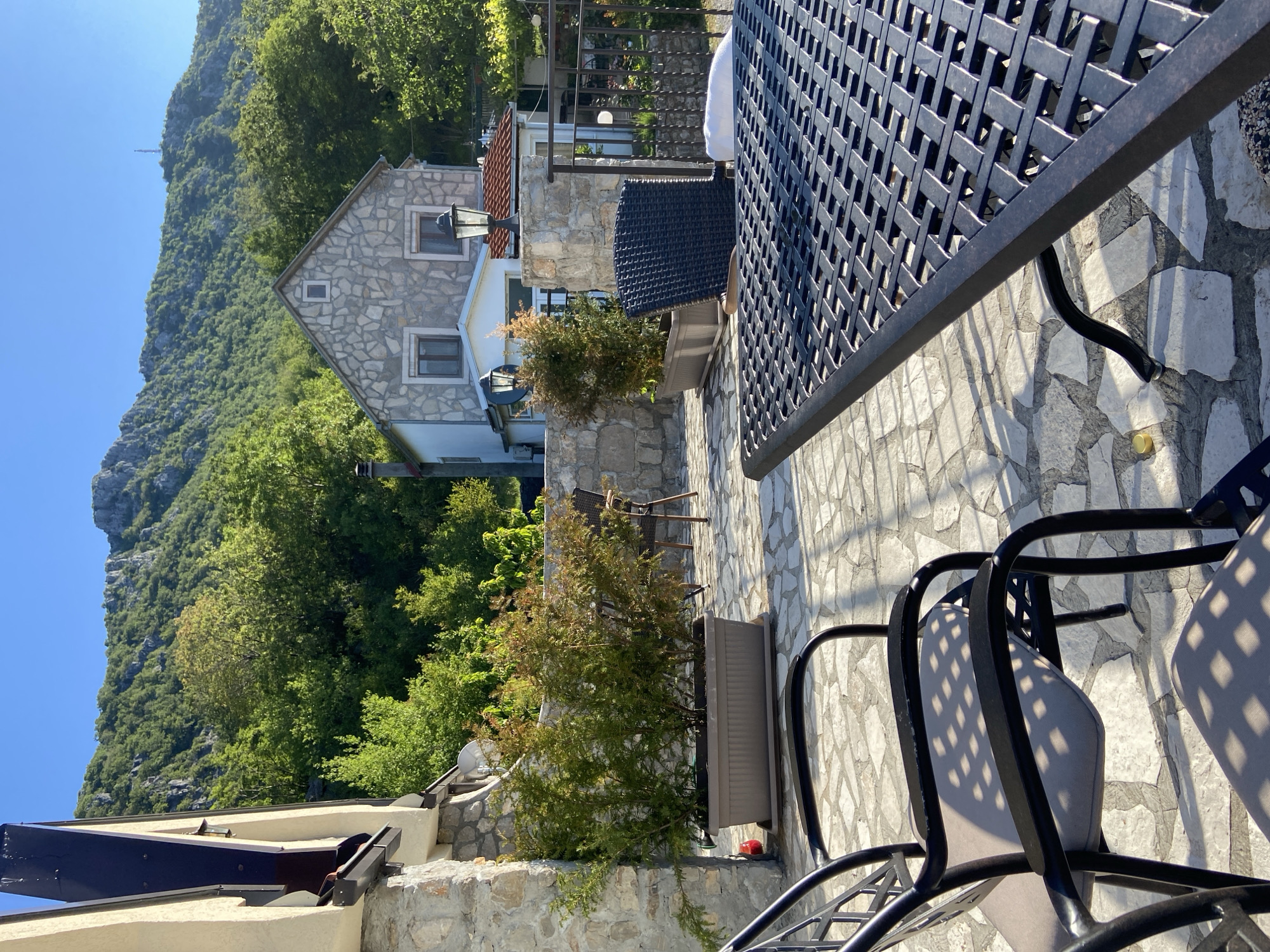 The image shows an outdoor patio in Sotonići, Montenegro. A stone house is visible in the background, nestled amongst trees and a mountain. In the foreground, there is a black metal table with a lattice design and matching chairs. A long rectangular planter with a green shrub sits along the edge of the patio. The patio itself is paved with light-colored stone. The scene is bathed in sunlight.