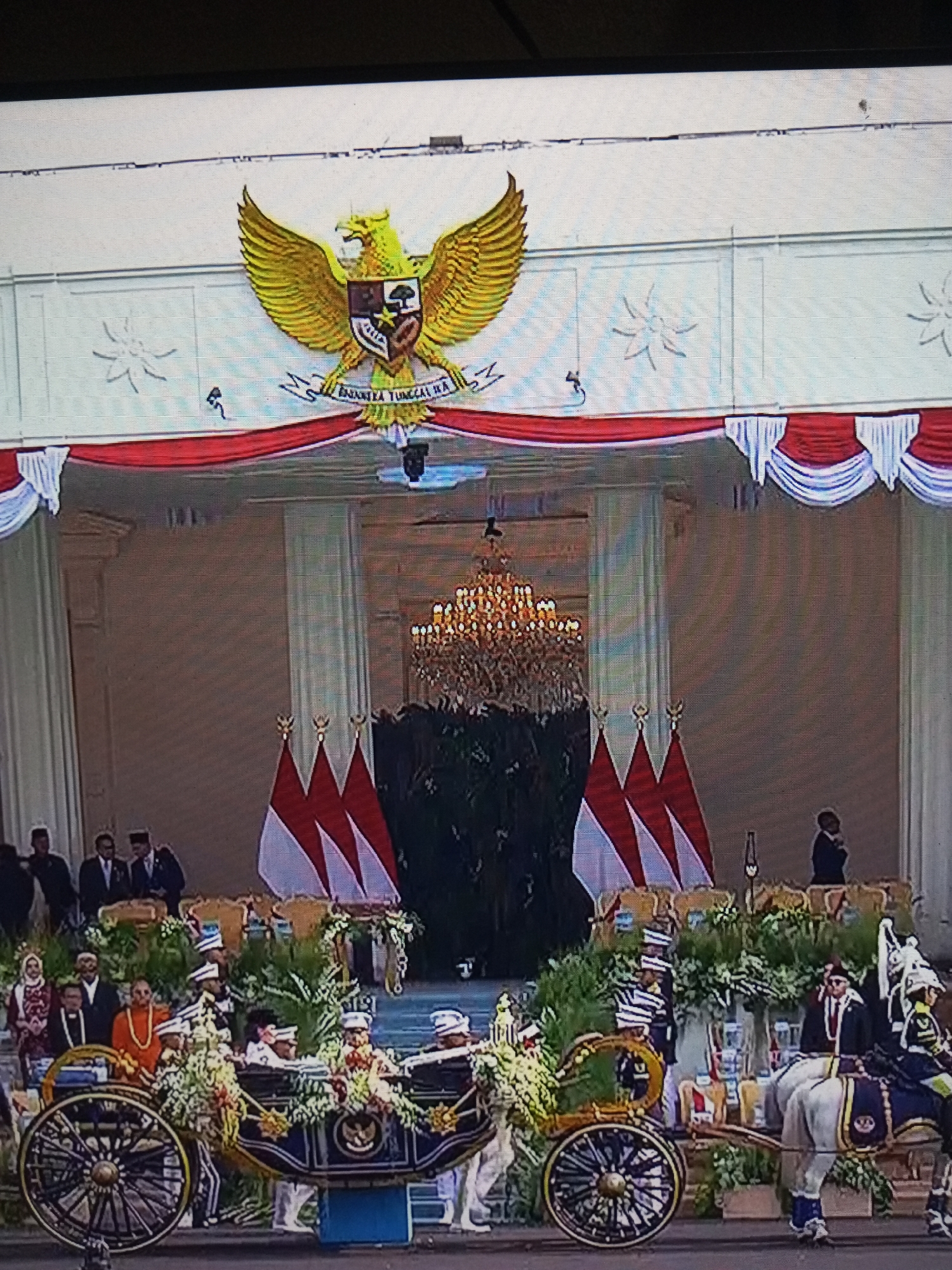 The image depicts a formal procession in Banyudono, Indonesia. In the foreground, a decorated horse-drawn carriage is being pulled by two white horses, suggesting a ceremonial event. The carriage is adorned with lush green foliage and floral arrangements, and several individuals are seated within it, appearing to be important figures. Uniformed personnel are present, some accompanying the carriage and others observing from the side. Behind the carriage, three Indonesian flags are displayed prominently. The backdrop features an ornate building with a large chandelier visible inside, indicating a significant architectural structure, possibly a palace or government building. A golden eagle emblem, the national symbol of Indonesia, is mounted above the building's facade. The overall scene conveys a sense of national pride and tradition.