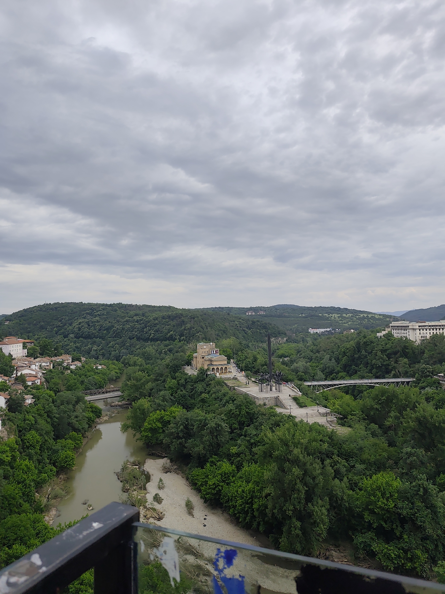 The image depicts a panoramic view of Veliko Tarnovo, Bulgaria, on an overcast day. A muddy river flows through a valley, with visible sandbanks and some vegetation. Dense green forests cover the surrounding hills. In the middle ground, a monument or public square with statues is visible. A bridge spans the river, connecting the different parts of the town. Residential buildings and other structures can be seen nestled among the trees. In the foreground, parts of a railing are visible, suggesting the photo was taken from an elevated viewpoint.