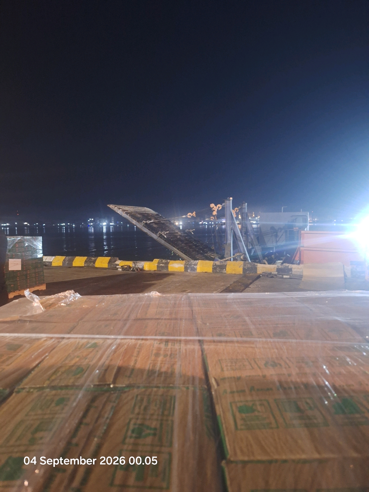 The image shows a nighttime scene at the Guntung River in Indonesia. In the foreground, a large stack of cardboard boxes wrapped in clear plastic is visible. The boxes appear to be uniform in size and have green markings on them. In the middle ground, a ramp or gangway extends from a vessel or structure towards the land. This ramp is marked with yellow and black hazard stripes. Beyond the ramp, there are lights from a city or industrial area reflecting on the water. The sky is dark, suggesting it is late at night or early morning. There are no people visible in the image.