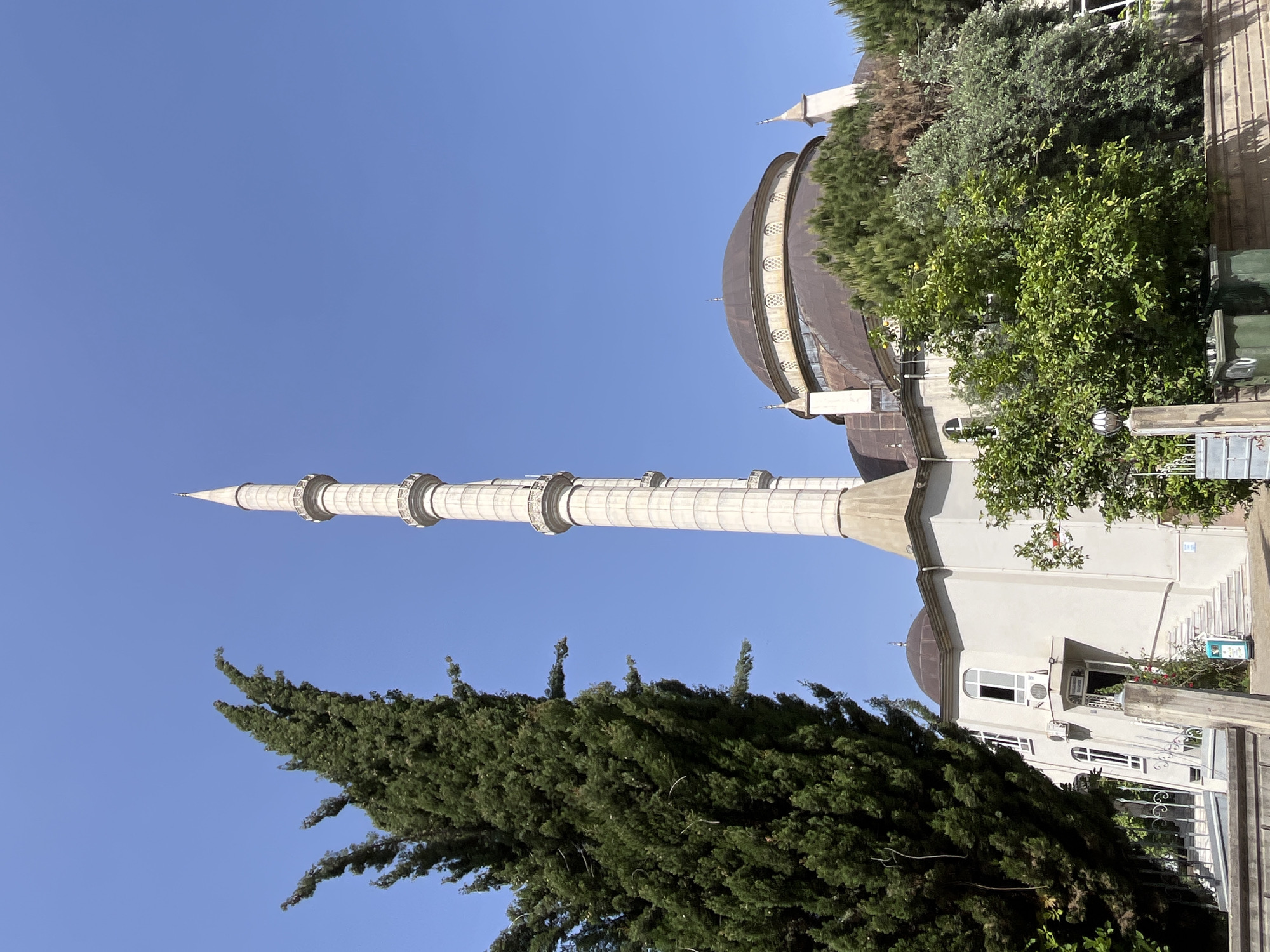 The image captures a mosque in Alanya, Turkey. The mosque features a tall, slender minaret with multiple cylindrical sections and a conical top, standing prominently against a clear blue sky. Adjacent to the minaret is the mosque's dome, covered in dark gray material. The mosque building is white, partially obscured by lush green trees. There is a small structure or building next to the mosque. The photograph is taken from a low angle, emphasizing the height of the minaret and the dome. The image is rotated 90 degrees to the left.