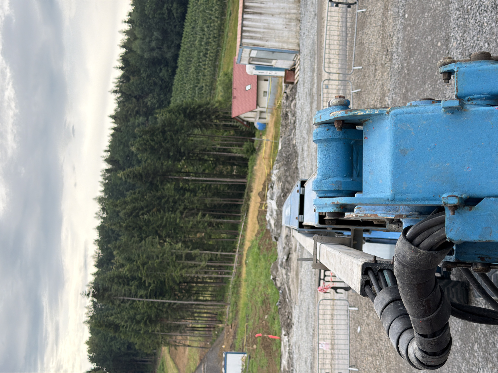 The image depicts a construction site in Stróża, Poland, on an overcast day. In the foreground, a close-up view of a blue construction machine is visible, with hydraulic hoses coiled around a metallic arm. Beyond the machine, a gravel-covered area stretches out, bordered by metal safety barriers. In the middle ground, a white construction trailer with a blue door and a building with a red roof are situated. Behind these structures, a dense forest of tall, green pine trees forms a backdrop. The ground slopes upwards towards the forest, with patches of grass and bare earth indicating ongoing construction activity. A dirt path or road is visible winding up the slope through the trees. The sky is filled with heavy, gray clouds. There are no people visible in the image.