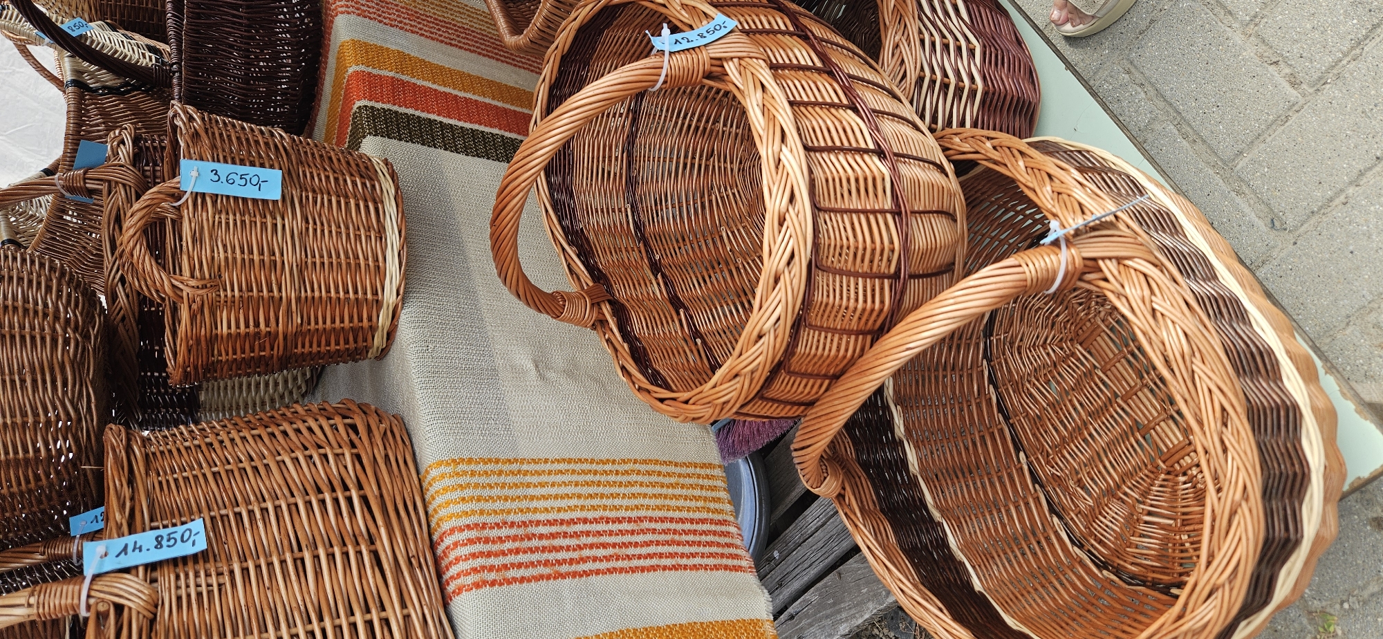 The image depicts an outdoor stall displaying an assortment of wicker baskets for sale. The baskets vary in size and design, with most being a warm, natural brown color, some featuring contrasting darker weaves. Several baskets have price tags attached with blue string. One tag clearly shows "3.650,-", another reads "4.850,-" and a third "12.850,-" on a blue tag with white lettering.
In the background, a striped fabric runner with bands of orange, yellow, and brown is spread across a table, on which some of the baskets are placed. A wooden surface is visible in the foreground, suggesting the stall is set up on a wooden deck or platform. The ground appears to be paved with concrete blocks, indicating an urban or semi-urban setting.
The image is taken from a slightly elevated angle, looking down at the baskets. No people are prominently featured, though a person's sandaled foot is partially visible on the right edge of the frame, indicating the presence of a shopper or vendor. The overall impression is one of a market or craft fair. Based on the provided information, this scene is located in the city of Hévíz, Hungary, likely at a market that sells local crafts.