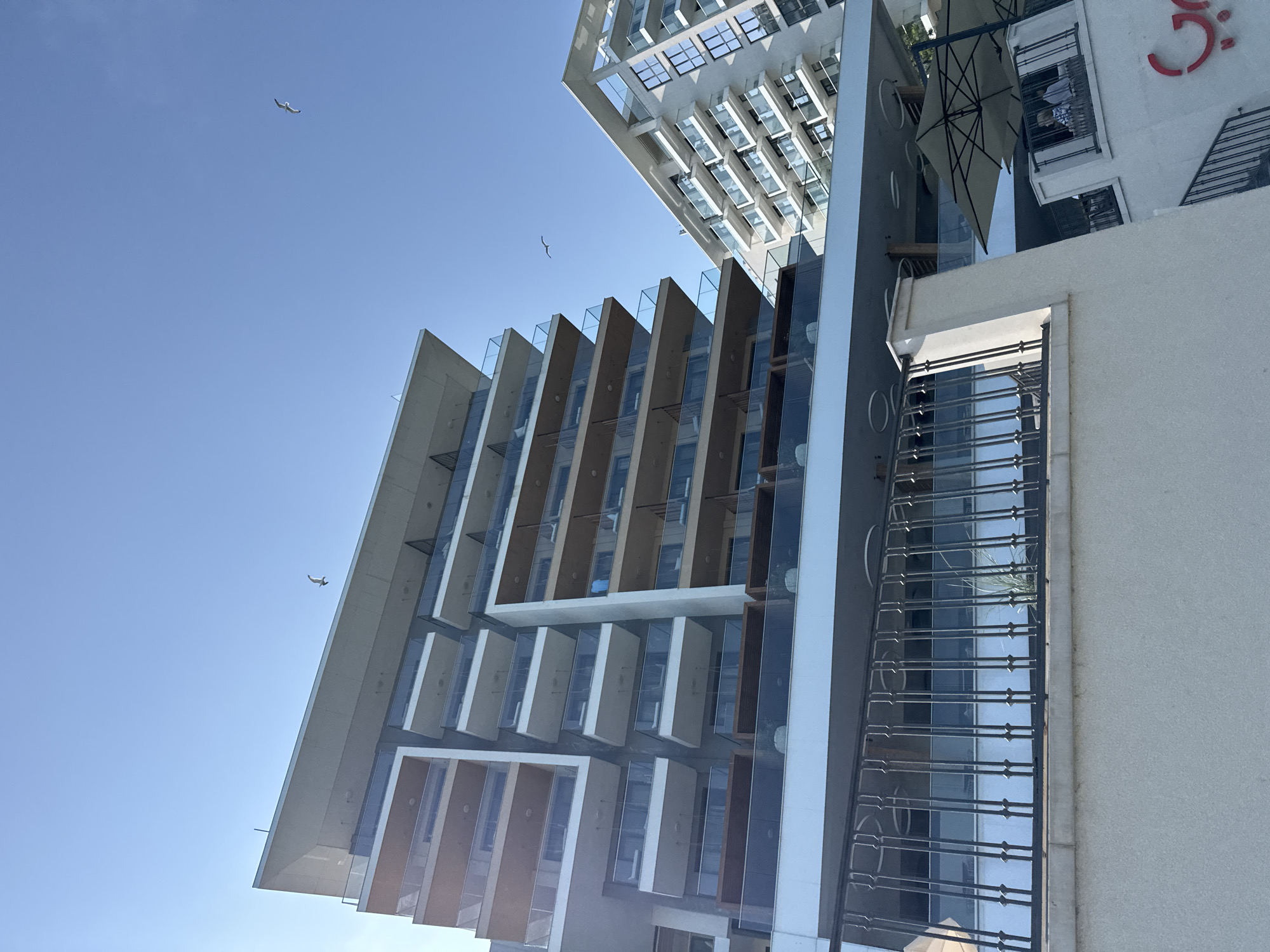 The image, when viewed in correct orientation, depicts a modern urban landscape under a clear, bright blue sky. A prominent contemporary building dominates the left and center, showcasing a stepped architectural design with angular balconies, extensive glass railings, and large windows. Its facade combines light-colored concrete elements with darker, possibly wooden or metallic, vertical panels. Partially visible at the top is another modern high-rise, characterized by a grid of numerous windows that reflect the sky.
On the right, a contrasting white building features a balcony protected by a decorative wrought-iron railing, with a glimpse of greenery behind it. Above this, a large, light-colored patio umbrella is unfurled, under which a person can be seen on a balcony. Red text or a logo, appearing to be in an Arabic-like script (resembling a stylized "ج"), is partially visible on the white wall above the umbrella.
Three birds, likely gulls, soar gracefully across the expansive blue sky. The scene is illuminated by clear sunlight, indicating a daytime setting with excellent weather conditions. The overall impression is one of modern city living within a serene, open environment.