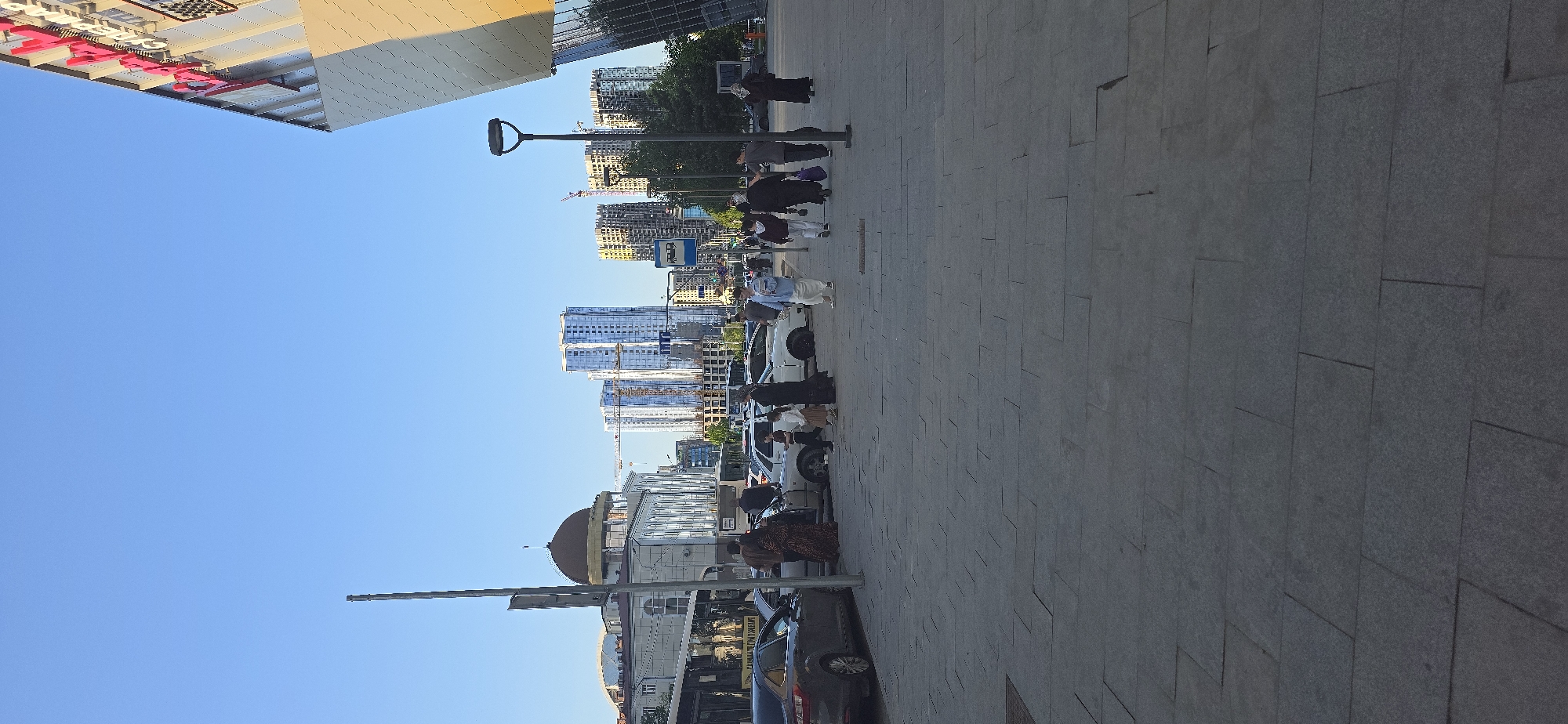 The image shows a street scene in Grozny, Russia. A group of people is walking along a sidewalk, with buildings and skyscrapers visible in the background. Cars are parked along the side of the street. The sky is clear and blue.