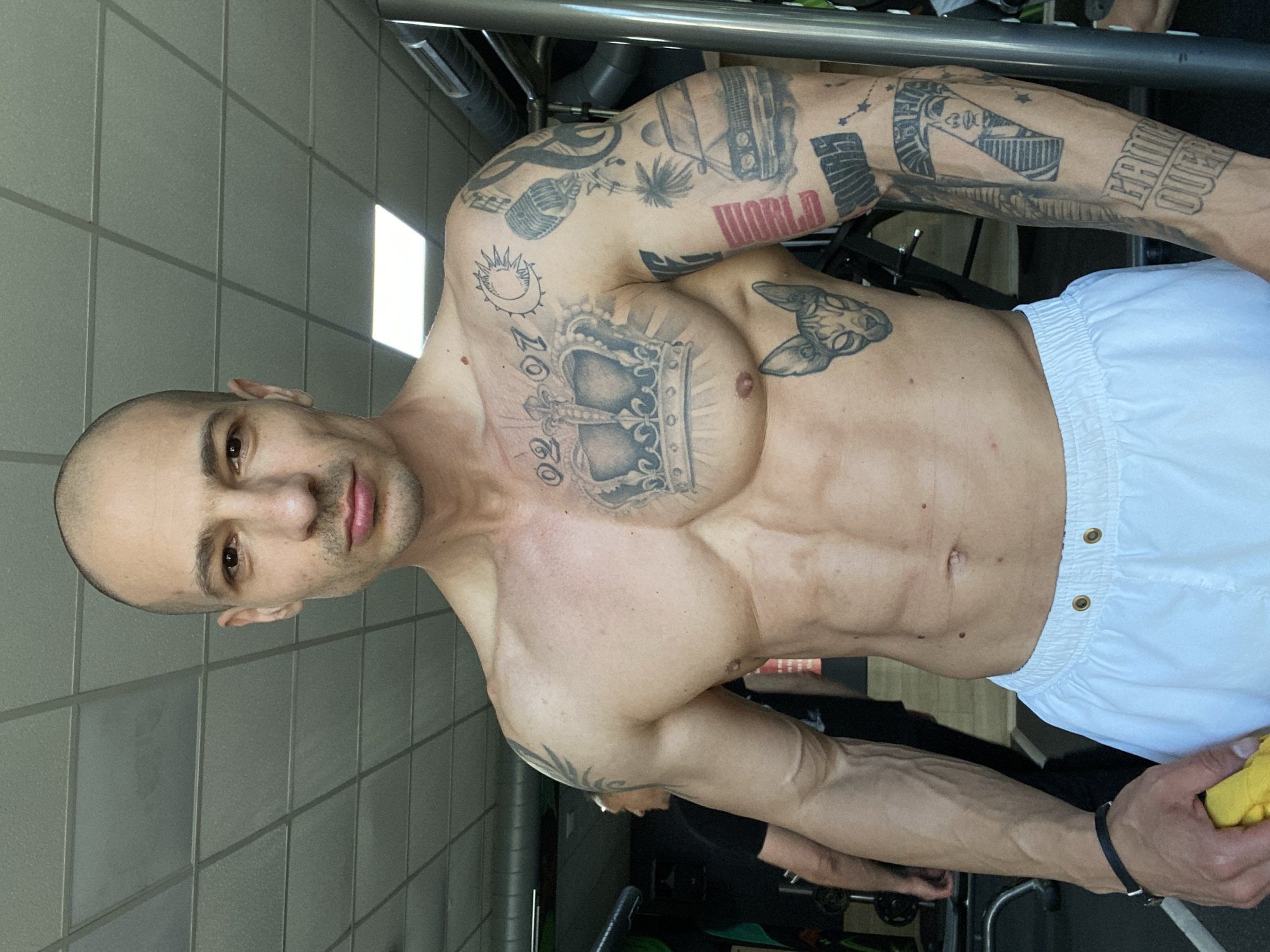 A shirtless, bald man with extensive tattoos stands in a room with light gray tiled walls and a gym-like setting. He wears white shorts. His tattoos cover his arms, chest, and upper body, depicting images such as a crown, a microphone, a sphinx, and the word "WORLD". The setting appears to be indoors, potentially a gym or exercise area, given the equipment visible in the background. The man holds a yellow object in his right hand. Pleven, Bulgaria, is not evident in the image.
