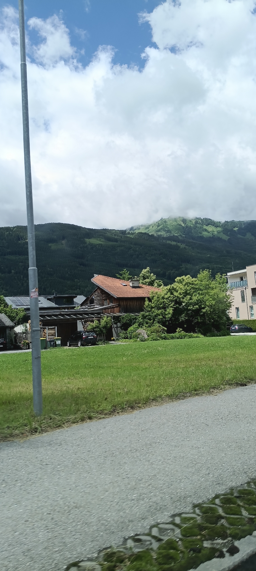 The image shows a residential area in Moos-Siedlung, Austria, with a focus on a traditional wooden house and surrounding landscape. In the foreground, a grey pole stands to the left, casting a shadow across a grassy lawn. Beyond the lawn, a traditional wooden house with a red tiled roof is visible, featuring a carport with a dark colored vehicle parked underneath. To the right of the house, a modern apartment building stands with a light-colored facade. In the background, a dense forest covers the lower slopes of a mountain range, with mist or clouds obscuring the higher peaks. The sky is partly cloudy, with patches of blue visible. The overall scene depicts a tranquil, rural setting.