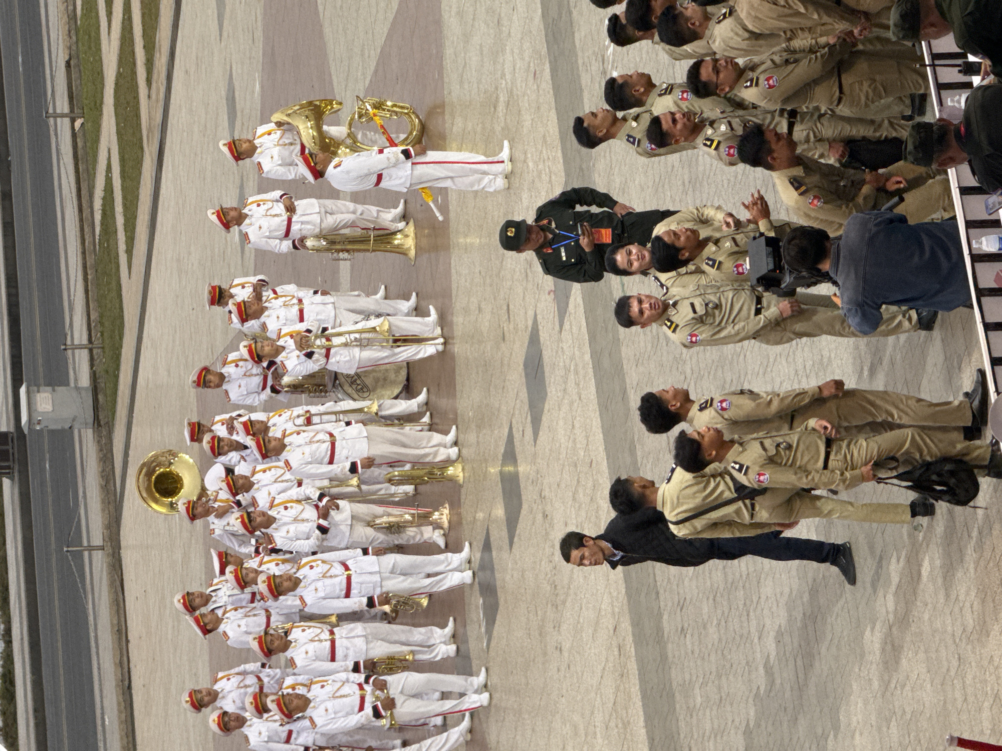 The image depicts a large group of people in uniform, many of whom are holding musical instruments, suggesting a band or orchestra. They are standing on a paved outdoor area that appears to be a public square or plaza. The architecture in the background is minimalist and modern, with a grey building and fencing visible. The people in uniform are predominantly male and are dressed in white and red outfits, with some wearing hats. Some individuals are also seen in the foreground in different uniforms, including a person in a dark green uniform who appears to be looking at a mobile phone, and others in tan uniforms. The overall scene conveys a sense of an organized event or ceremony. The image does not provide any specific visual cues to indicate the location is Da Lat, Vietnam.