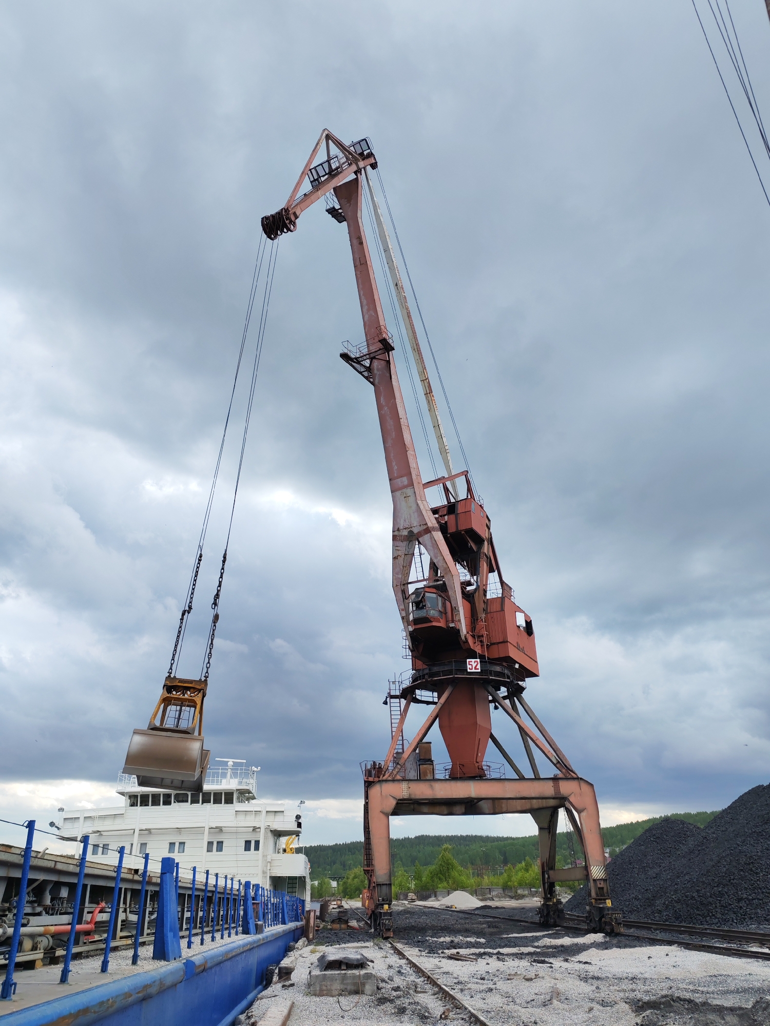 The image depicts an active industrial port scene in Medvezhyegorsk, Russia, under a heavily overcast sky. The primary subject is a large, reddish-orange gantry crane, exhibiting signs of wear and heavy use, positioned centrally. Its imposing structure features multiple levels and operator cabins, with the number "52" visible on its lower section.
Suspended from the crane's cables is a heavy-duty excavator bucket or grabber, colored brown/gold. This grabber is positioned directly above the deck of a large, white ship or vessel, indicating ongoing or imminent bulk cargo handling operations, such as loading or unloading. The ship, with its multi-story white superstructure and blue deck railings, is docked alongside a pier.
On the ground to the right of the crane, large piles of black, granular material, consistent with coal or other bulk commodities, are visible. Multiple rail tracks run beneath the crane's gantry base and along the waterfront, suggesting railway transport integration within the port's logistics. In the distance, a dense green forest covers rolling hills, contrasting with the industrial foreground.
The weather is characterized by a thick, gray, cloudy sky, suggesting an overcast daytime. No individuals are visible in the frame. The scene conveys a sense of large-scale industrial activity and transportation of bulk goods in a northern, waterfront setting.