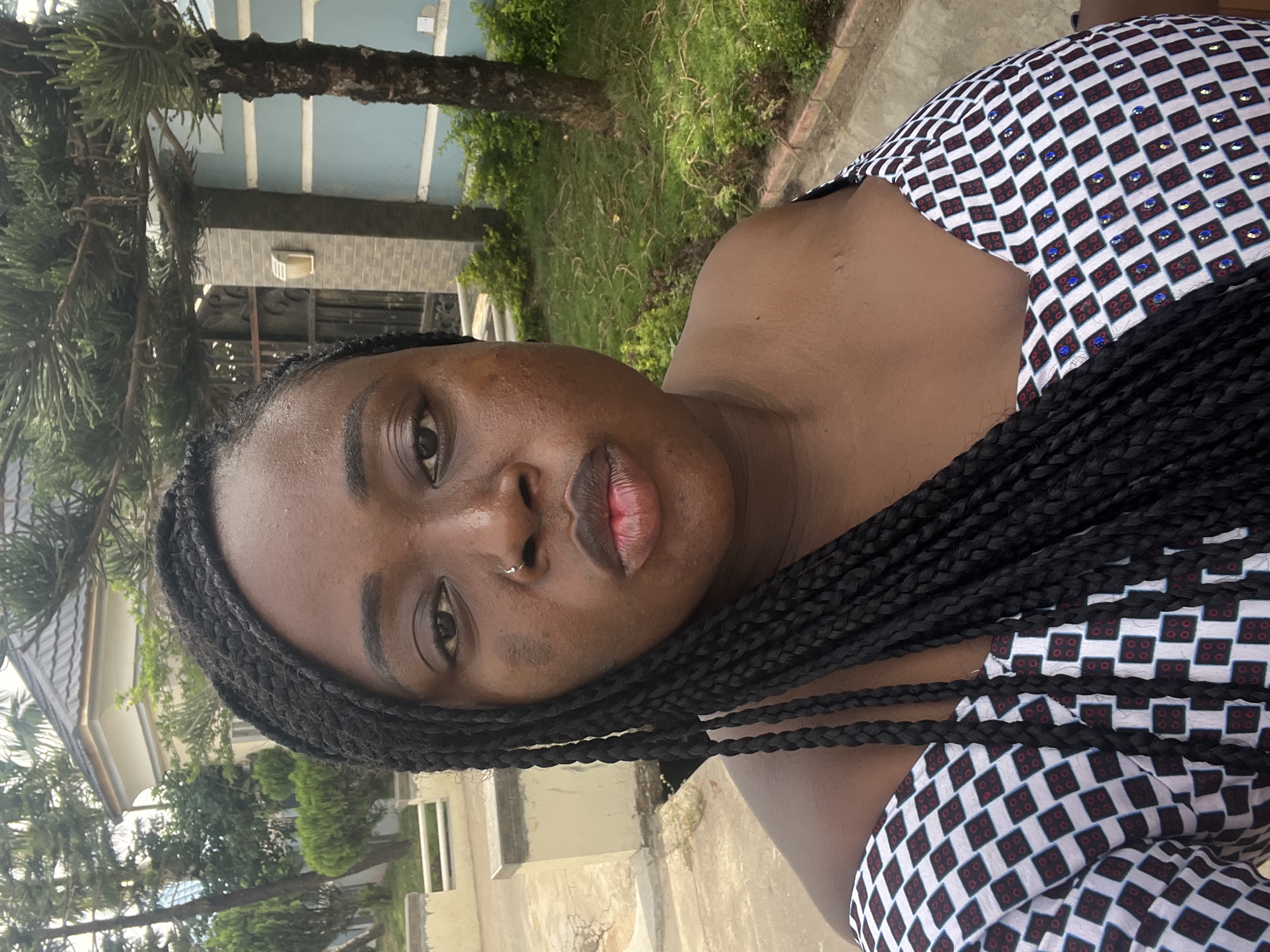 The image depicts a dark-skinned woman with braided hair, wearing a checkered patterned top with embellished details. She has a nose ring. The background includes greenery, a tree trunk, and sections of buildings with pale blue and beige coloring. The photo appears to be taken outdoors in daylight. The exact location within Abuja, Nigeria, cannot be determined from the image alone.