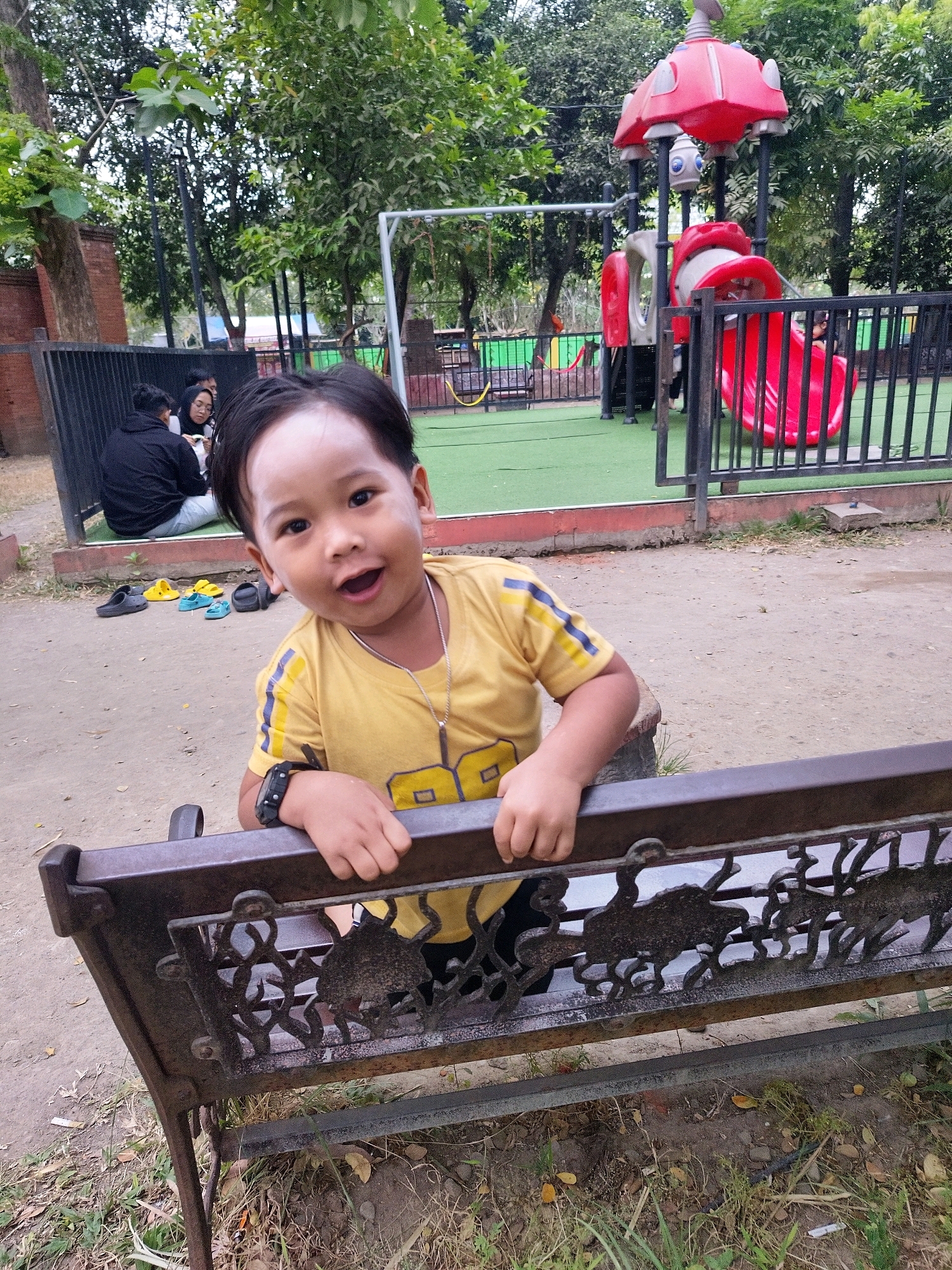 A young boy, appearing to be around three to five years old, is leaning over a dark metal bench with ornate, cutout designs. He is wearing a yellow t-shirt with blue and yellow stripes on the sleeves and a silver necklace. The boy has dark hair and is smiling broadly, with his mouth open and his eyes looking directly at the camera. His hands are resting on the bench. Behind him, to the left, two adults are sitting on the ground behind a black metal fence, with one person wearing a black hoodie and the other wearing a headscarf. In the background, there is a red and gray playground structure with a slide, and more trees and foliage. Several pairs of colorful sandals are scattered on the dirt ground in front of the bench. The overall setting appears to be an outdoor recreational area in Mojokerto, Indonesia.