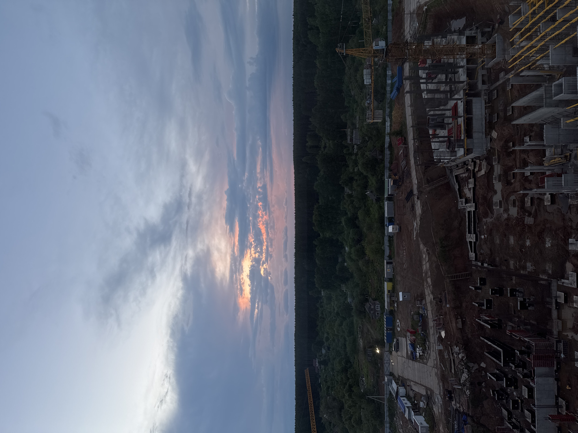 This is a photograph of a construction site at dusk in Izhevsk, Russia. The sky is filled with dramatic clouds, with a band of soft orange and pink light near the horizon, suggesting the last rays of the setting sun. In the foreground, a large construction site is visible, with concrete foundations and structural elements of buildings in various stages of completion. Construction cranes, one prominently yellow, are positioned on the site. Beyond the construction area, there is a dense forest line, and further in the distance, scattered buildings and infrastructure are discernible. The overall impression is one of ongoing development within a natural landscape.
