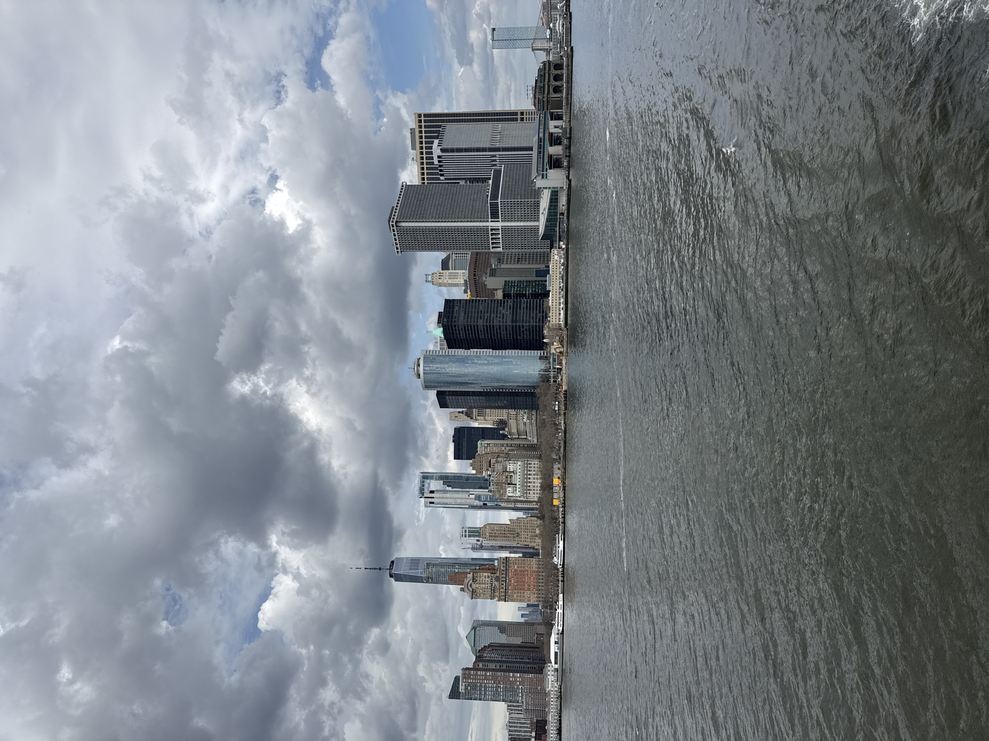 This image shows the New York City skyline. The skyscrapers of lower Manhattan are visible, including One World Trade Center. The water in the foreground is likely part of the Hudson or East River. The sky is cloudy with patches of blue.