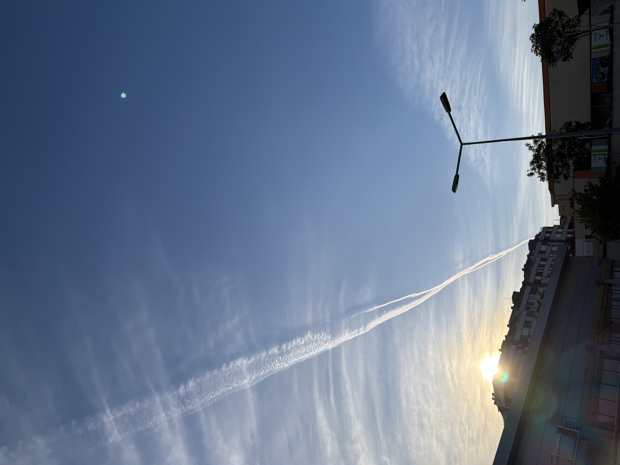 The image shows a low-angle view of the sky in what appears to be an urban environment. In the top right corner, there are buildings. A Y-shaped street lamp stands prominently against the sky. A prominent contrail stretches across the sky from the bottom right of the image, near where the sun is positioned. There are light, wispy clouds throughout the blue sky. The sun is shining brightly and creating a lens flare effect. Based on the architecture and signage in the image, the location may be in Burgas, Bulgaria.