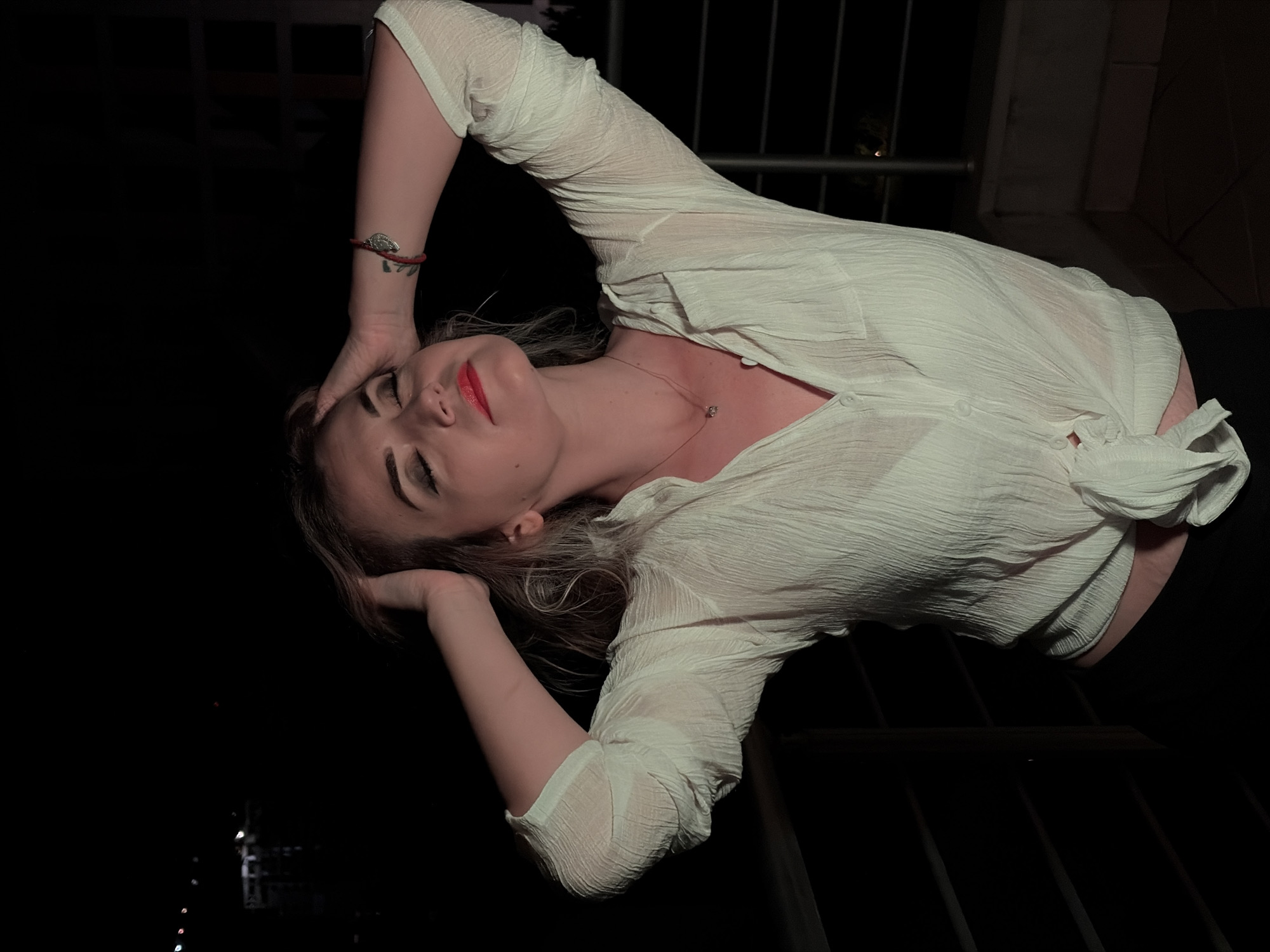 An adult female is depicted from the mid-thighs up, posing outdoors at night on what appears to be a balcony in Budva, Montenegro. She has light-colored hair, closed eyes, and red lipstick. Both hands are raised to her head, with fingers interlocked within her hair, and her elbows are bent and extended outwards. Her head is tilted slightly back and to the viewer's left. She wears a light-colored, textured, long-sleeved button-up shirt, which is tied at the waist, revealing a small portion of her abdomen. A lighter-colored undergarment is visible beneath the shirt's somewhat sheer fabric. Dark trousers or a skirt are partially visible below the shirt's knot. Accessories include a thin chain necklace with a small pendant, a red string bracelet with a silver-toned leaf charm on her right wrist, and a small tattoo on her right forearm. The scene is illuminated by an artificial light source from the front, highlighting the subject against a predominantly dark background. Distant, blurred artificial lights, consistent with a city or coastal town at night, are visible on the horizon. A metallic balcony railing with horizontal bars is present in the foreground and mid-ground.
