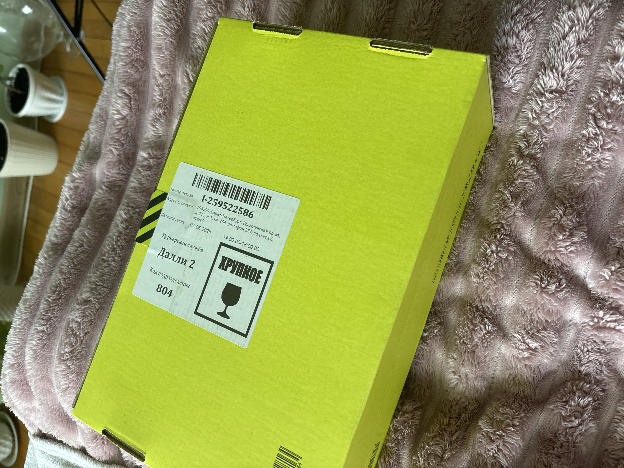 An unboxing scene is captured in this top-down, medium shot image, featuring a bright yellow cardboard box partially obscuring a soft, pink, fuzzy blanket. The box is angled towards the viewer from the left side of the frame. A white label with black text and a barcode is affixed to the top of the box. Text on the label indicates the location as "Novoe Devyatkino, Russia" and includes details like "Courier Service," "Order Number," and delivery information. A graphic of a wine glass within a square frame is also present on the label, suggesting the contents are fragile. The background shows a wooden floor with several potted plants.