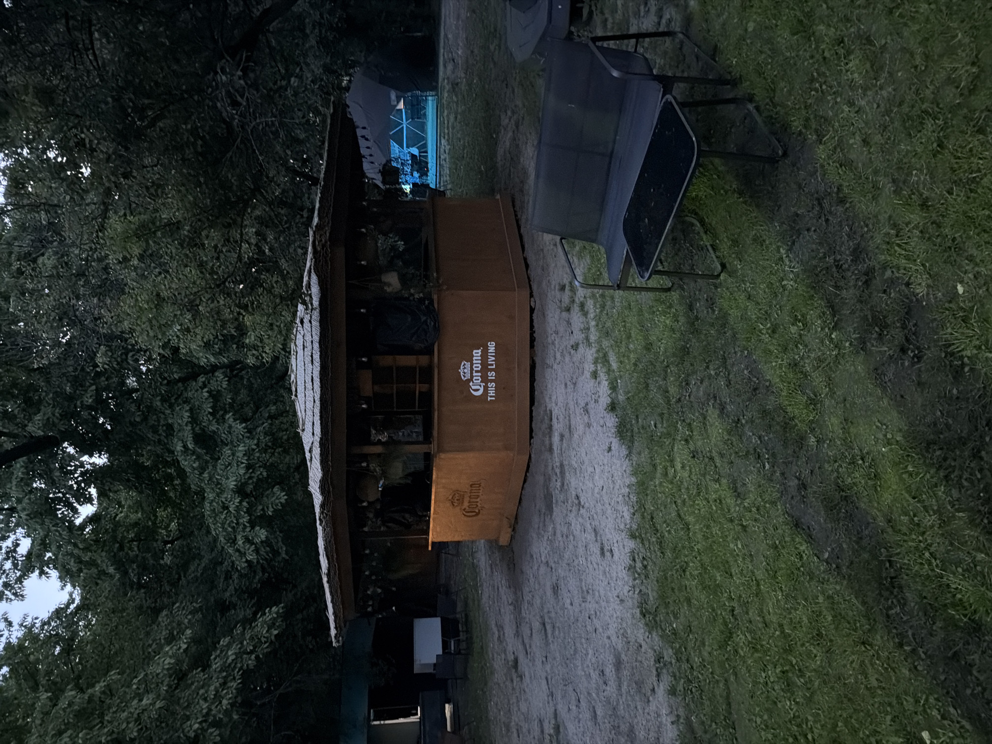 This image captures an outdoor bar area, situated in Kyiv, Ukraine, under the conditions of dusk or nighttime. A centrally located, multi-sided wooden bar structure, possibly octagonal or hexagonal, features a prominent thatched roof with string lights suspended beneath its eaves. The bar's exterior displays "Corona" branding with the tagline "THIS IS LIVING" visible on at least two distinct panels. The interior or serving sections of the bar appear dark, suggesting it is either closed or unlit.
The immediate ground around the bar consists of packed dirt and sparse vegetation, transitioning to a more defined grassy area in the foreground. The entire scene is enveloped by dense, dark-foliaged trees, indicating a park or wooded setting, with a dark blue-grey sky visible through the tree canopy. To the left, behind the bar, a light-colored object, resembling a cooler, and several dark chairs are visible adjacent to a dark, possibly building-like structure with an open entrance. To the right, a large, dark geodesic dome tent is distinctly illuminated from within by a vivid blue light.
In the foreground, on a patch of grass, a dark two-seater bench is paired with a rectangular dark table. No individuals or animals are present in the frame, and no specific actions or interactions are depicted. The lighting is low, relying on ambient twilight and the artificial illumination from the bar and the dome structure.