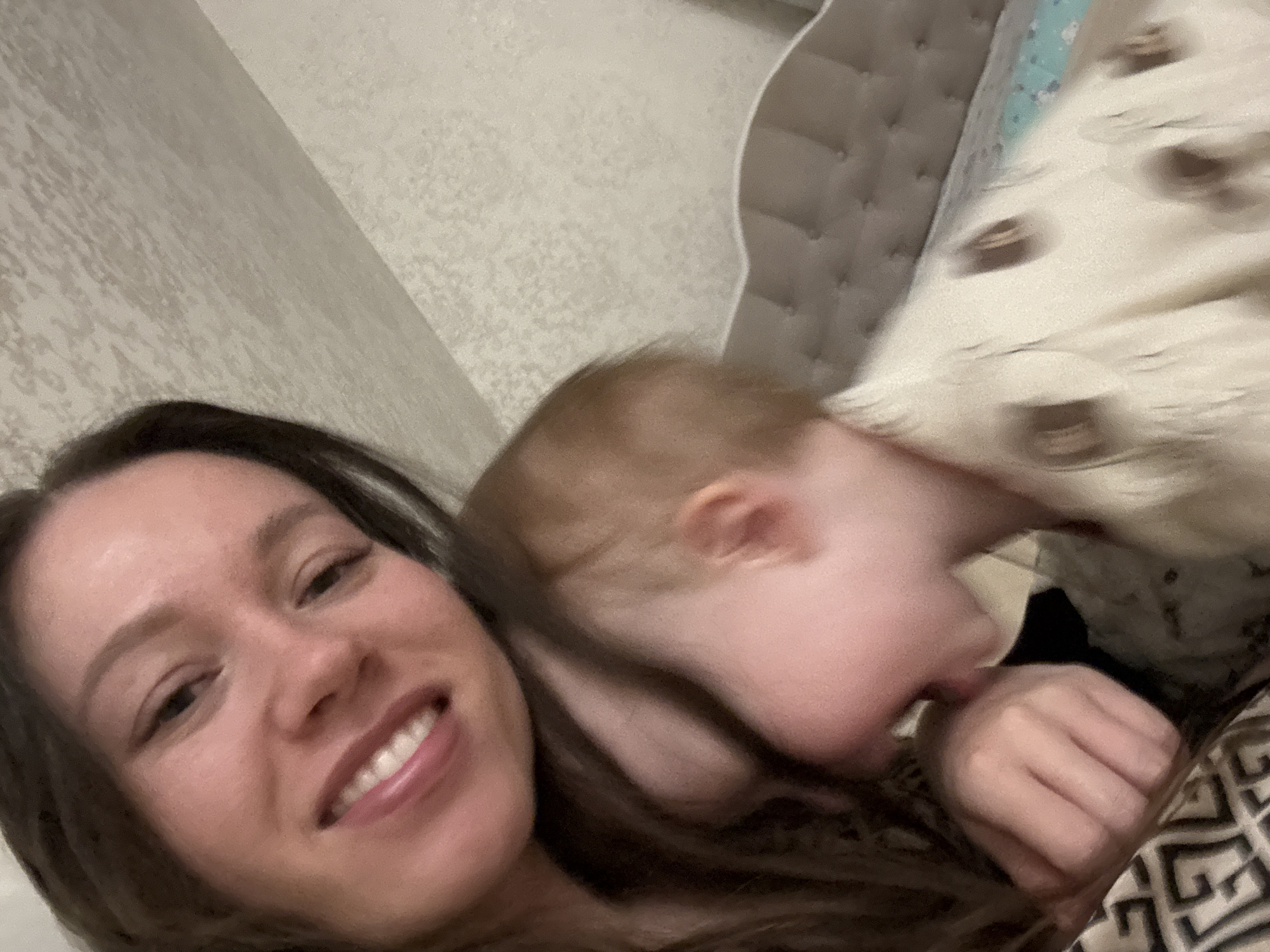 An indoor close-up photograph depicts an adult female and an infant or toddler. The scene is described as being in the city of Ферма, Russia.
The adult female, with long dark brown hair and fair skin, smiles towards the camera, her upper teeth visible. She occupies the upper-left portion of the frame. Her left hand, partially covered by dark patterned fabric, rests on her chest or shoulder in the lower-left foreground. An infant or toddler, with light brown hair, leans into the adult's left side. Its profile is visible, with its mouth slightly open. The child wears a light-colored garment featuring faint dark circular or irregular patterns.
The background consists of a light-colored, subtly textured wall. On the right side, a bed is visible with a light-colored, tufted headboard. The bedding includes a light-colored blanket with faint dark patterns and a thin turquoise border along its edge.