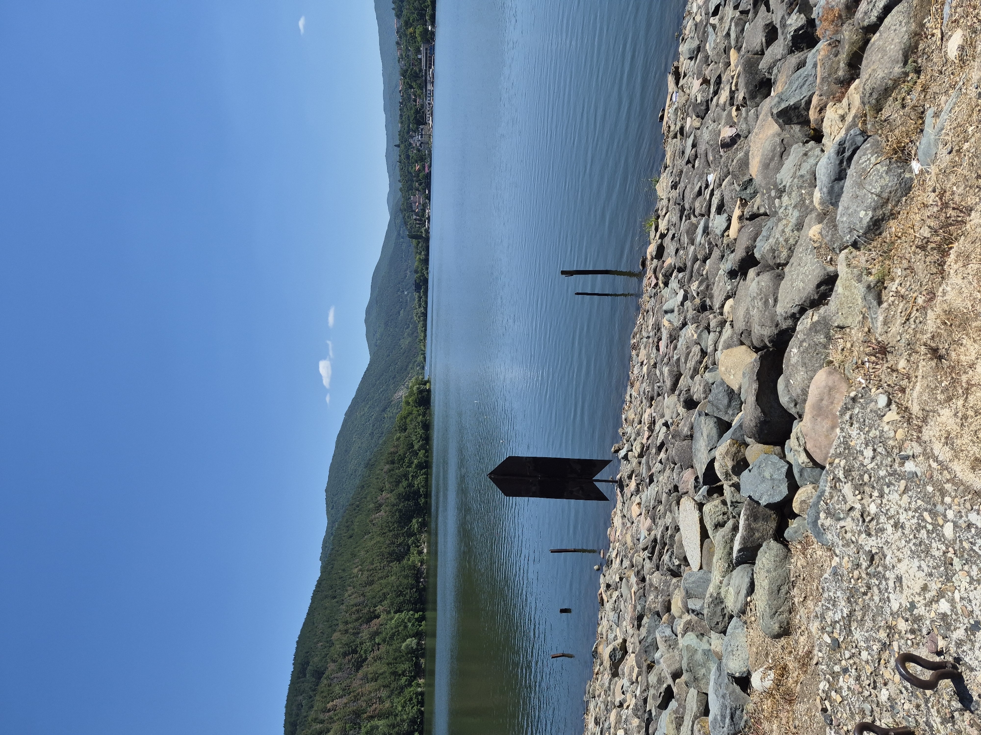 The image depicts a scenic view of Pancharevo Lake in Pancharevo, Bulgaria, under a clear blue sky. The foreground is dominated by a rocky shoreline, composed of numerous grey and brown boulders of varying sizes. A few dry grasses and weeds are scattered among the rocks. In the middle ground, a dark, arrow-shaped marker stands in the water, accompanied by several wooden posts. The lake itself is calm and reflects the surrounding landscape, with gentle ripples on its surface. The background features rolling green hills covered in dense forest, and a distant town or village with houses and buildings nestled among trees along the water's edge. The overall scene conveys a peaceful and natural environment.