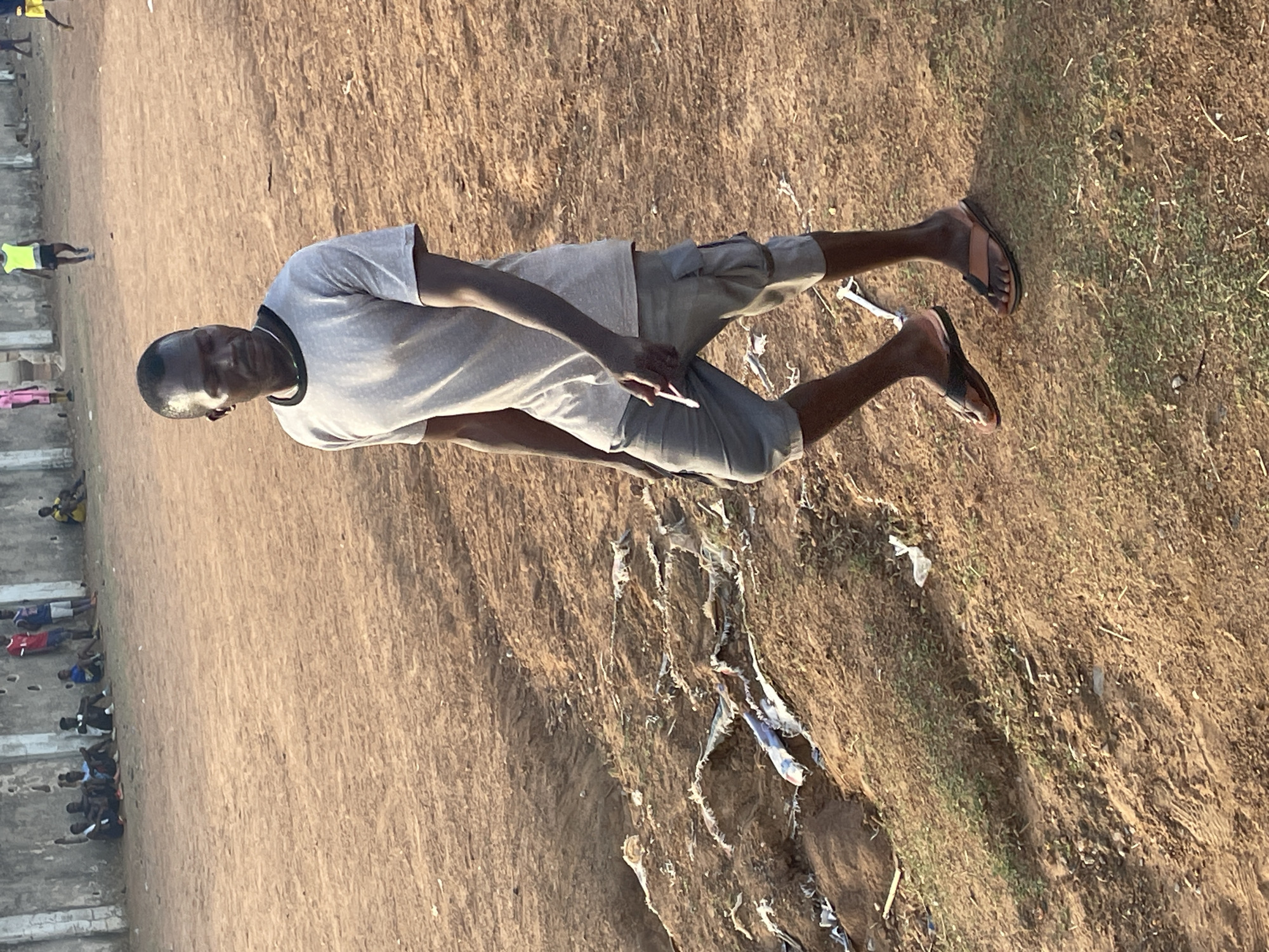 The image shows an individual standing on a dirt and grassy area adjacent to stadium seating. The person is wearing a light-colored t-shirt, gray shorts, and sandals. The ground is a mix of dirt and sparse vegetation, with scattered debris. Other individuals are visible in the stadium seating. The location is Akwanga, Nigeria.