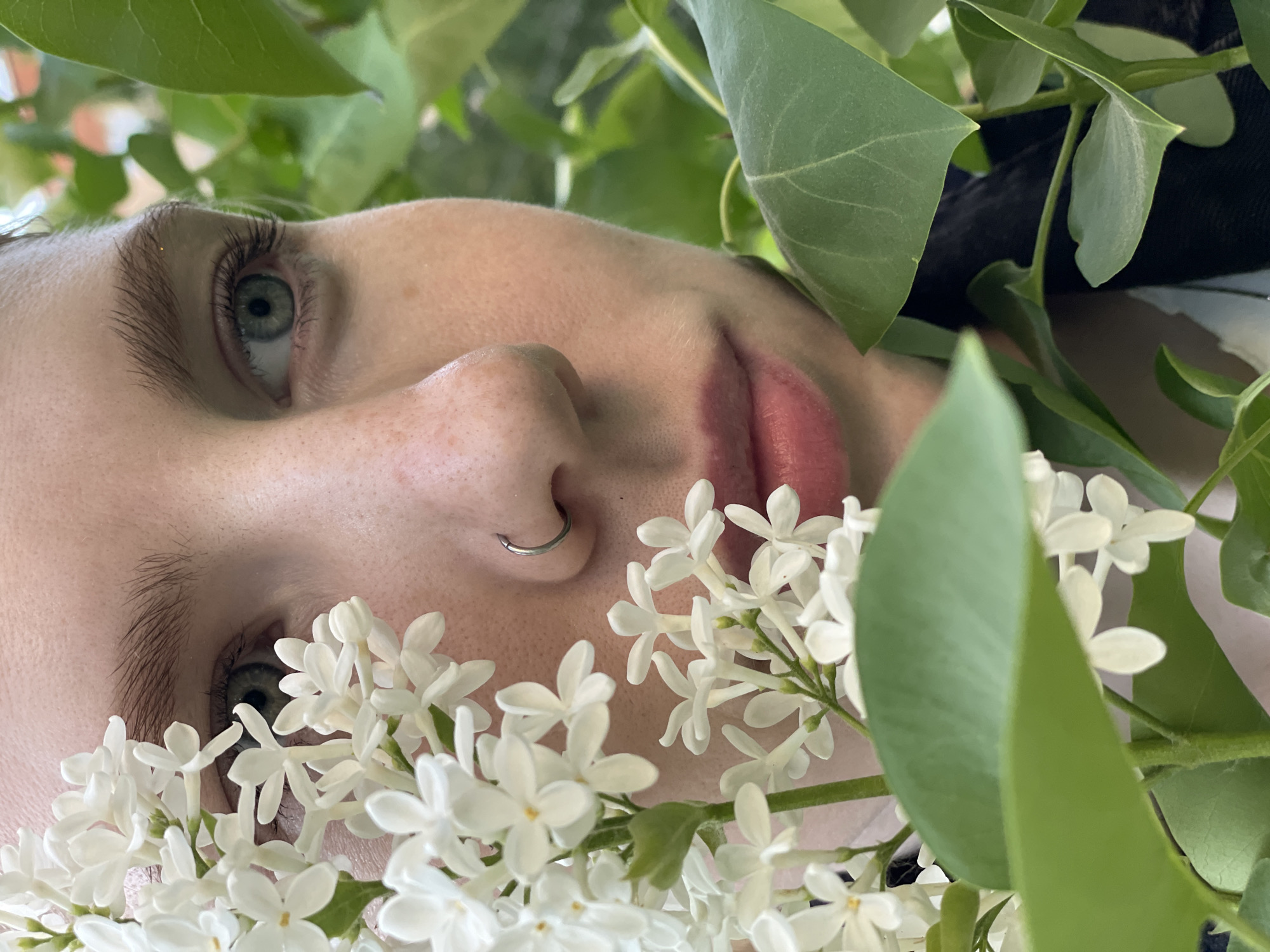 A close-up, angled portrait shot focuses on a person's face surrounded by greenery and white flowers. The person, with light skin, freckles, and short hair, has green eyes and a silver nose ring. Their lips are tinted pink, and they appear to be lying down. The face is framed by green leaves and clusters of small white flowers, likely lilacs, which partially obscure the lower half of the face. Some black clothing is visible behind the leaves at the top right. The overall impression is fresh and floral. The location is not discernible from the image.