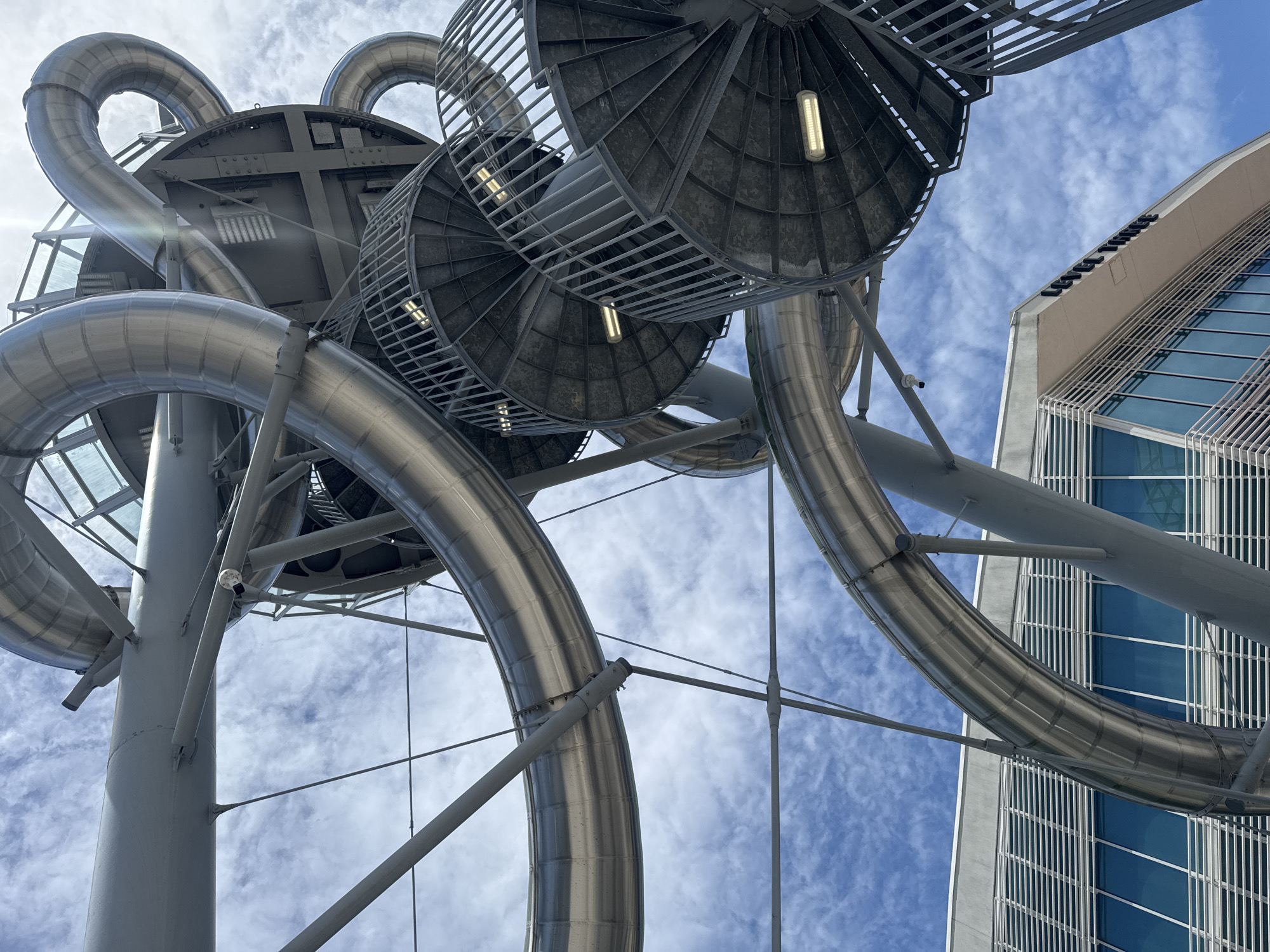 A low-angle outdoor photograph captures a large, modern, metallic spiral slide structure against a cloudy blue sky. The slide is made of polished metal tubes that curve and twist in complex formations, supported by a grey steel framework. Multiple circular, metal spiral staircases with open railings are visible, integrated into the structure, with industrial lights illuminating the steps.
Below the slide structure, the upper section of a contemporary building is visible. This building features a light-colored facade with prominent horizontal louvers and large, blue-tinted glass windows. Text is visible on the building's exterior, reading "LEVEL THREE". The scene appears to be in the city of Aventura, United States of America. No people are present in the frame.