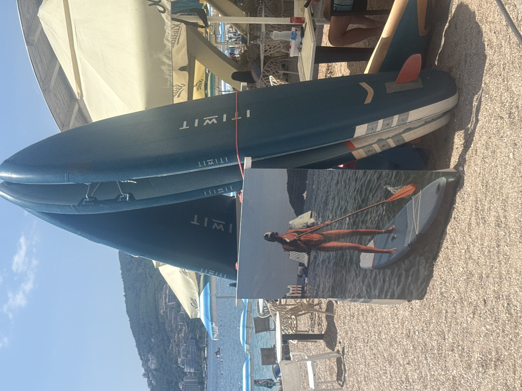 This media file depicts a scene at a beach in Budva, Montenegro. The primary subjects are several stand-up paddleboards (SUPs) stacked on top of each other. The SUPs are dark blue with white accents and feature red fins. One of the SUPs has "ITIWIT" printed on its side.
In the foreground, a photograph is placed on the sand, showing a person in a bathing suit standing on a SUP in the water, holding a paddle. The person is wearing a hat and goggles. The background of this photograph shows a body of water and a beach with chairs and umbrellas.
The larger scene shows the SUPs positioned on a sandy beach, with a view of the sea and a hilly coastline in the distance, dotted with buildings. This coastline is characteristic of Budva. The sky is a clear blue, suggesting a sunny day, likely during daylight hours, perhaps late morning or afternoon given the shadow lengths. The overall impression is of a leisure or recreational setting at a seaside resort. There is no discernible emotion conveyed by the subjects in the main image, other than the implied activity of a beach vacation.